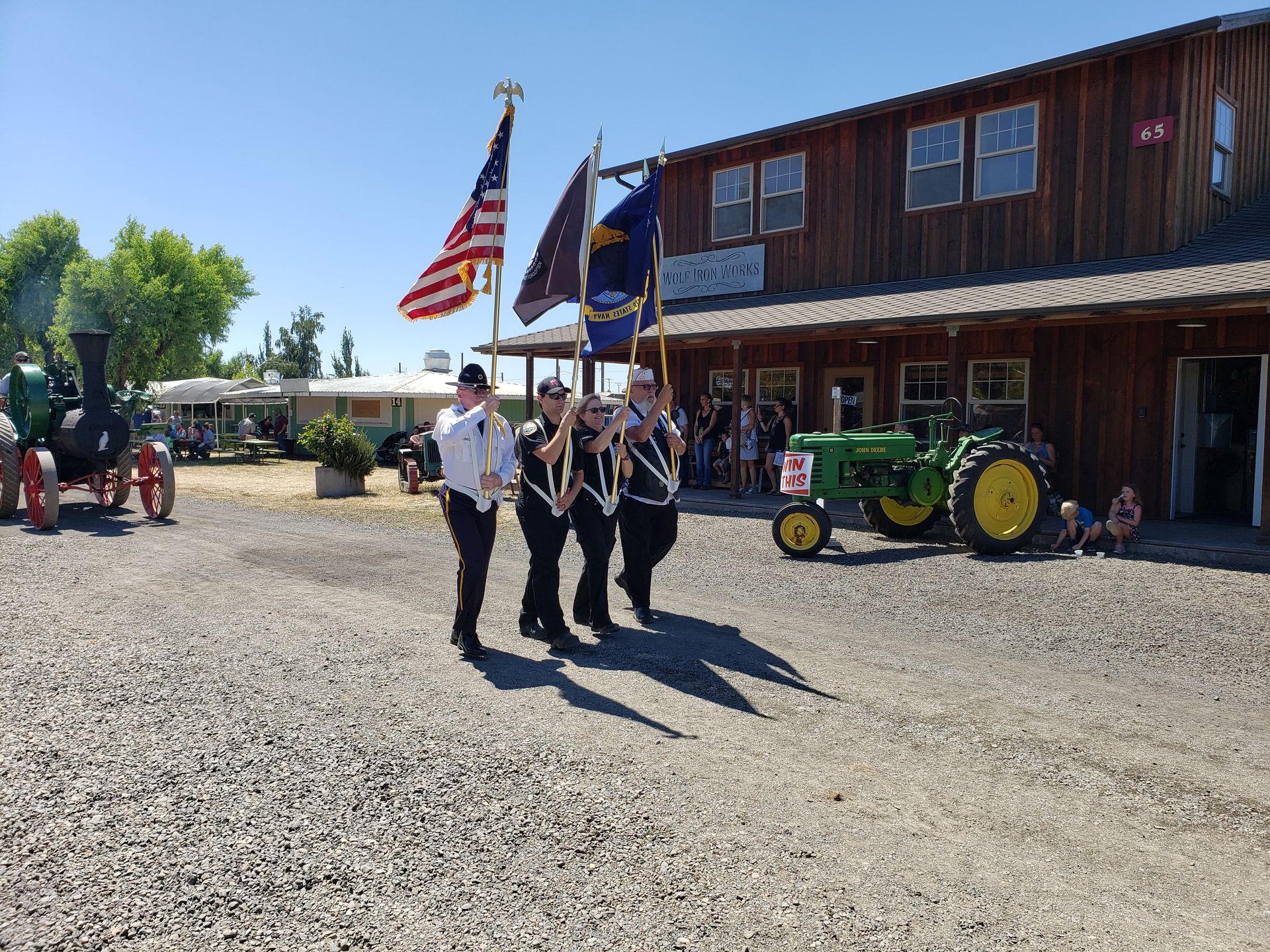 Color guard marching with flags in front of a rustic building and parked tractors on a sunny day