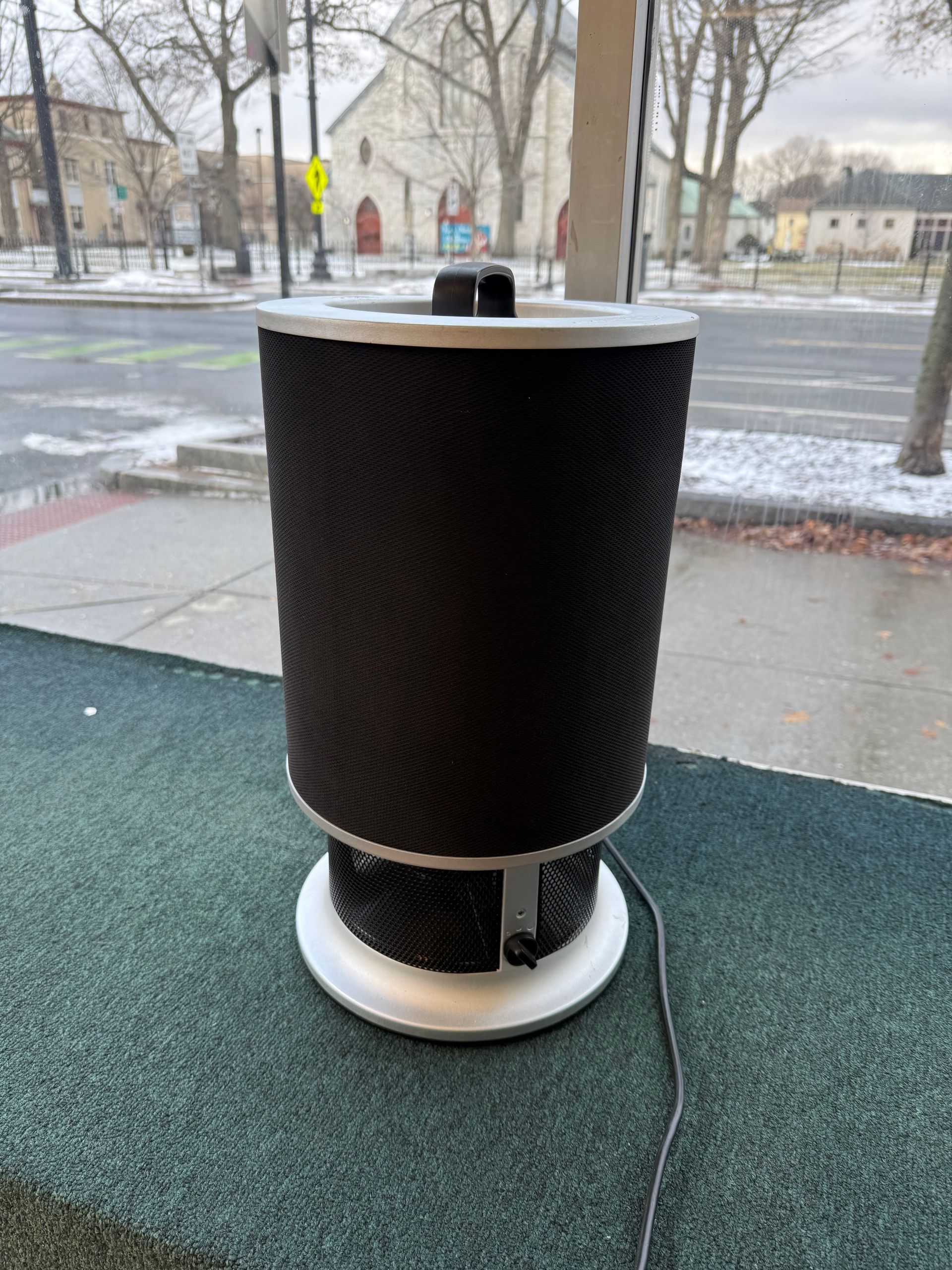 A tall, cylindrical air purifier with a black mesh exterior and silver base, sitting on a green surface near a window.