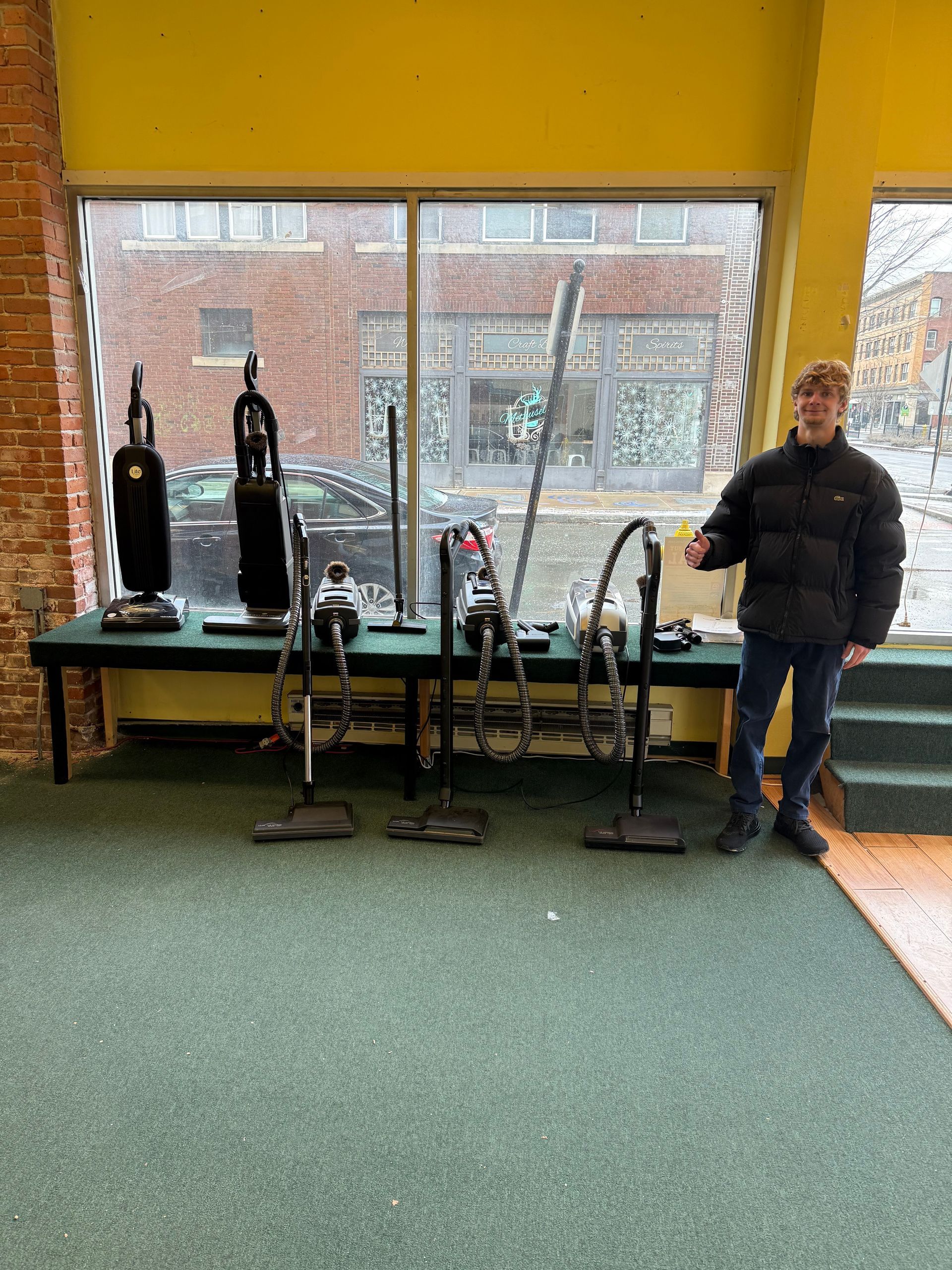 A person stands next to a table displaying several vacuum cleaners inside a room with large windows overlooking a street.