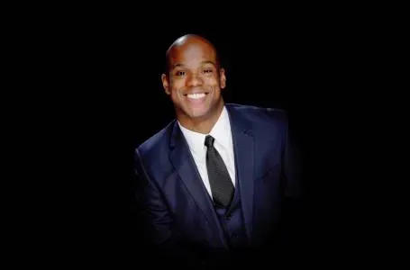 Man in blue suit smiles brightly against a black background.