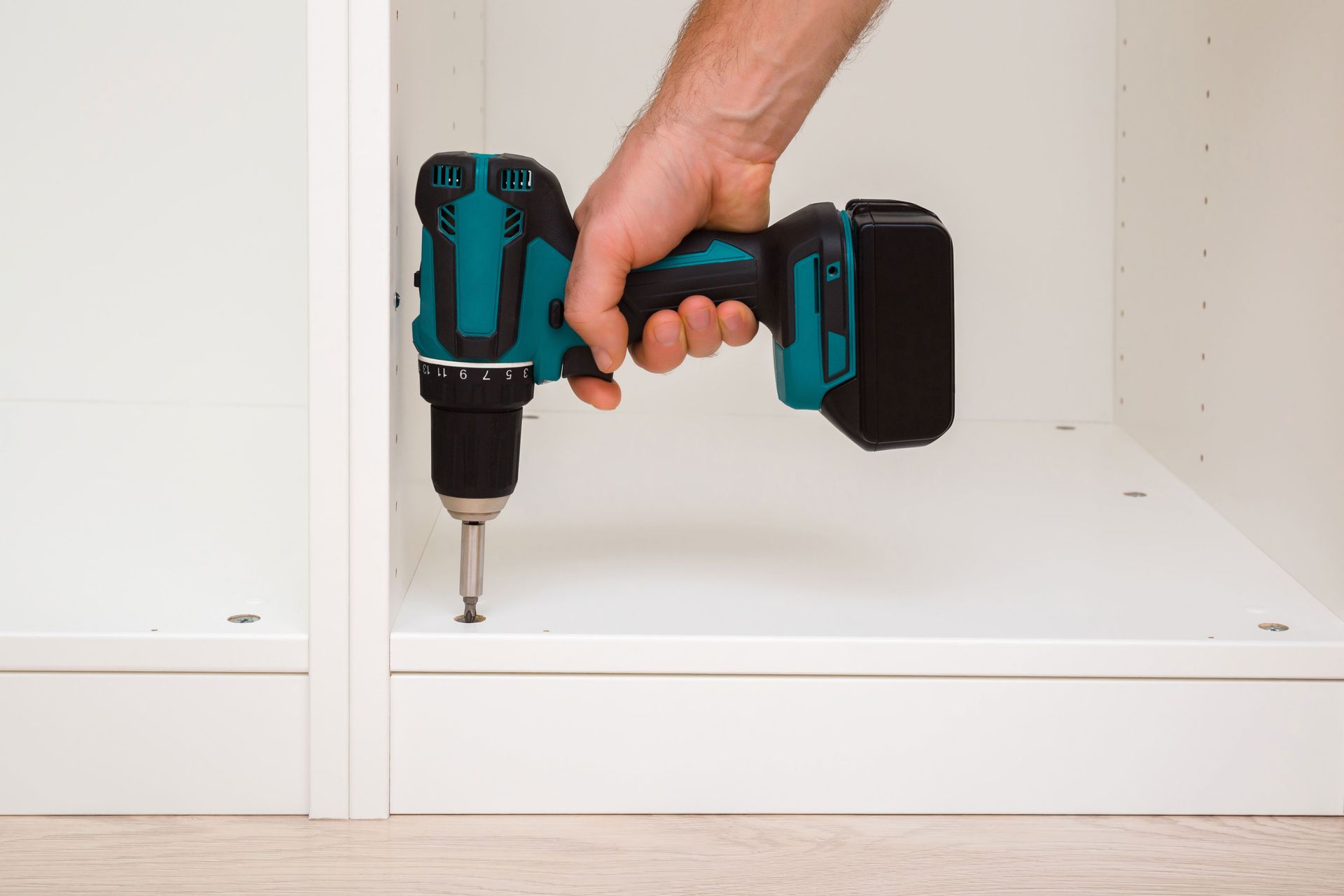 Hand holding a teal cordless drill to install a screw inside a white cabinet.