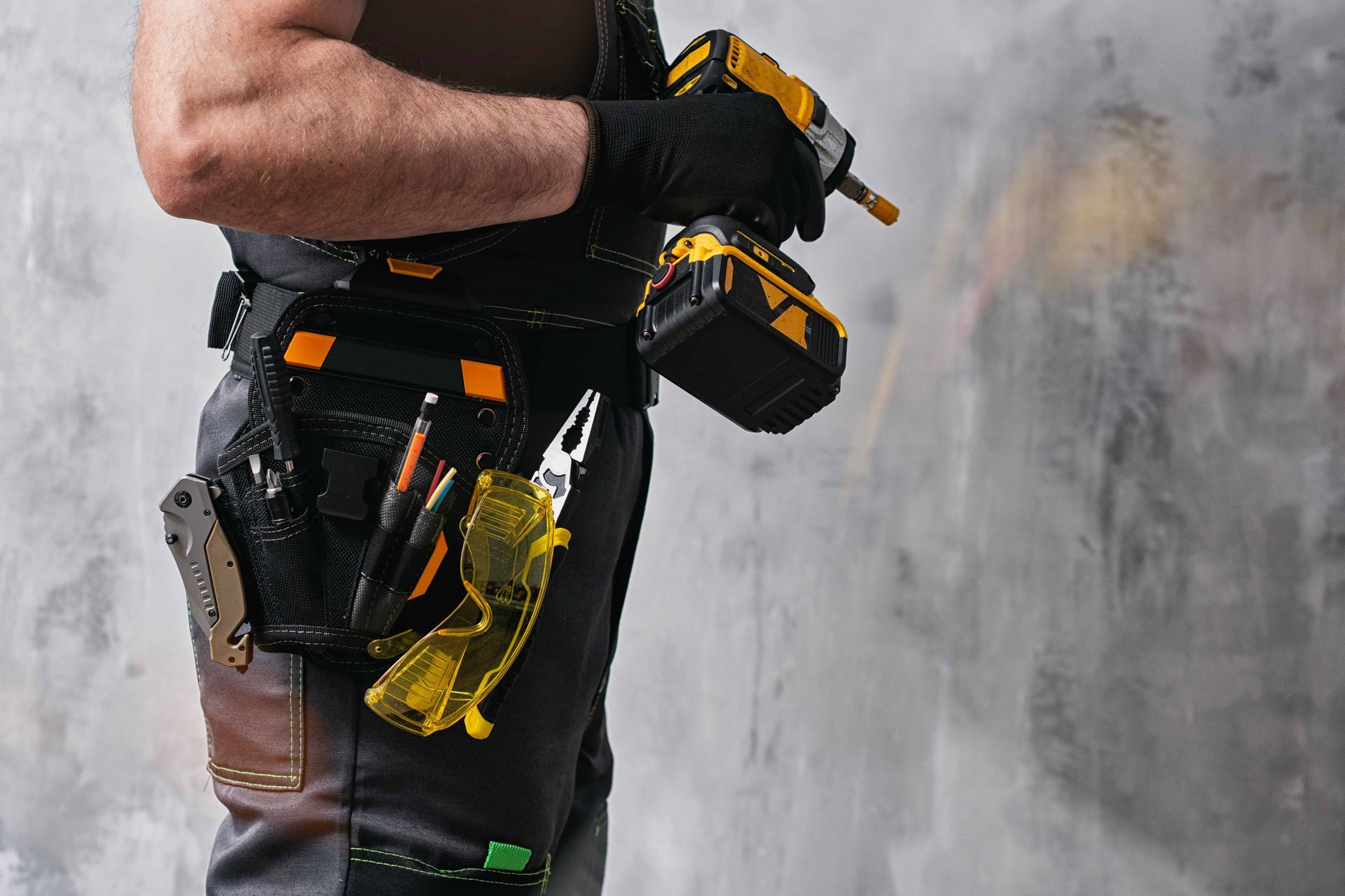 Close-up of a worker’s tool belt with black and yellow tools against a gray wall.