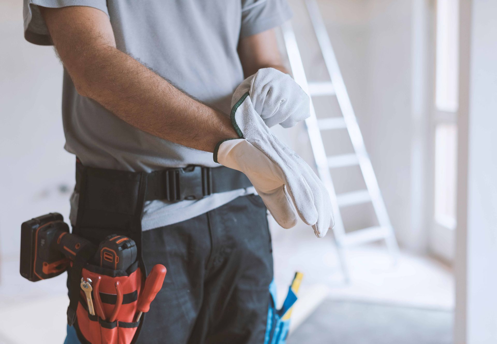 Worker holding a pair of white gloves at a construction site, with tools in a belt and a ladder in the background