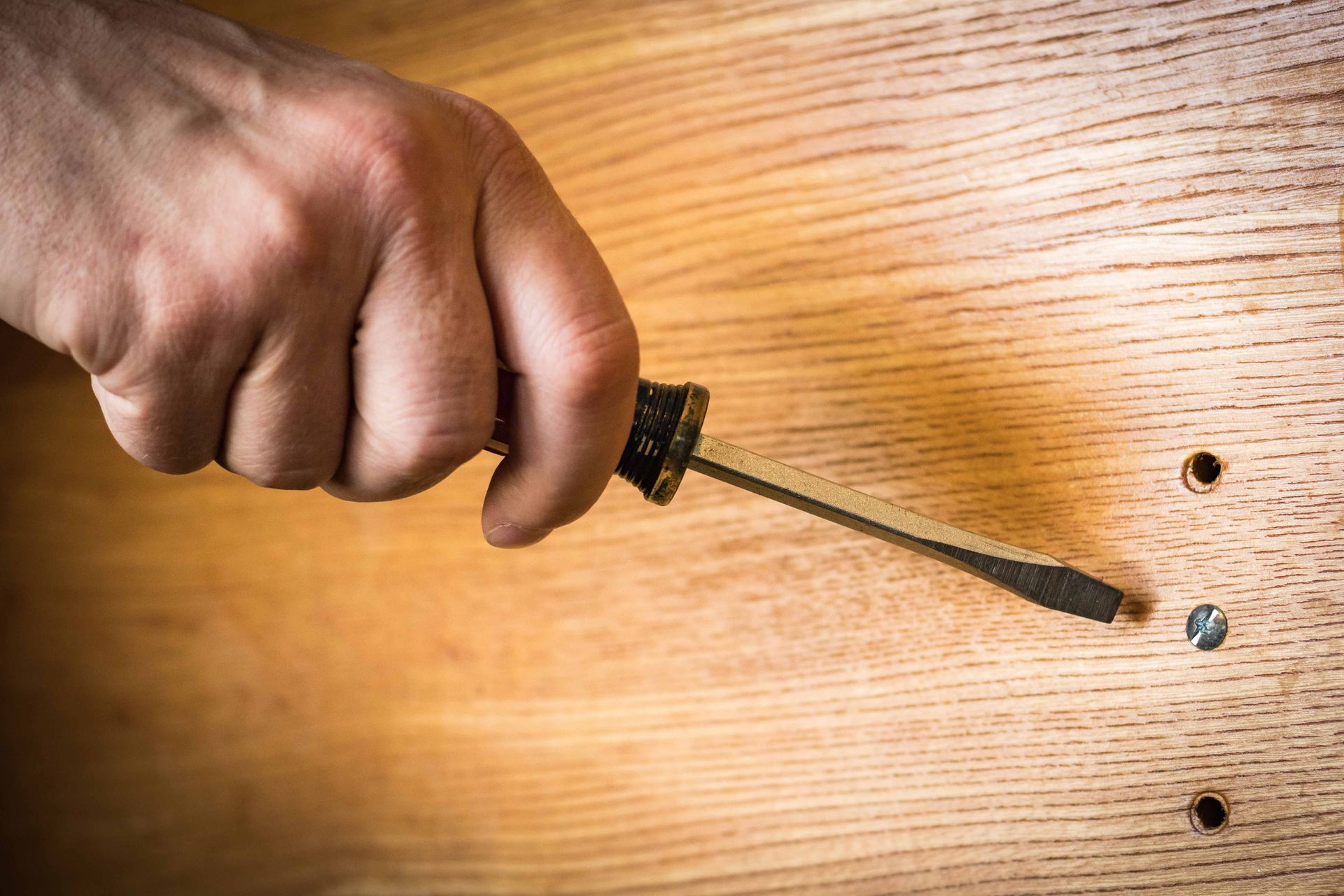 Hand holding a screwdriver near screws on a wooden surface