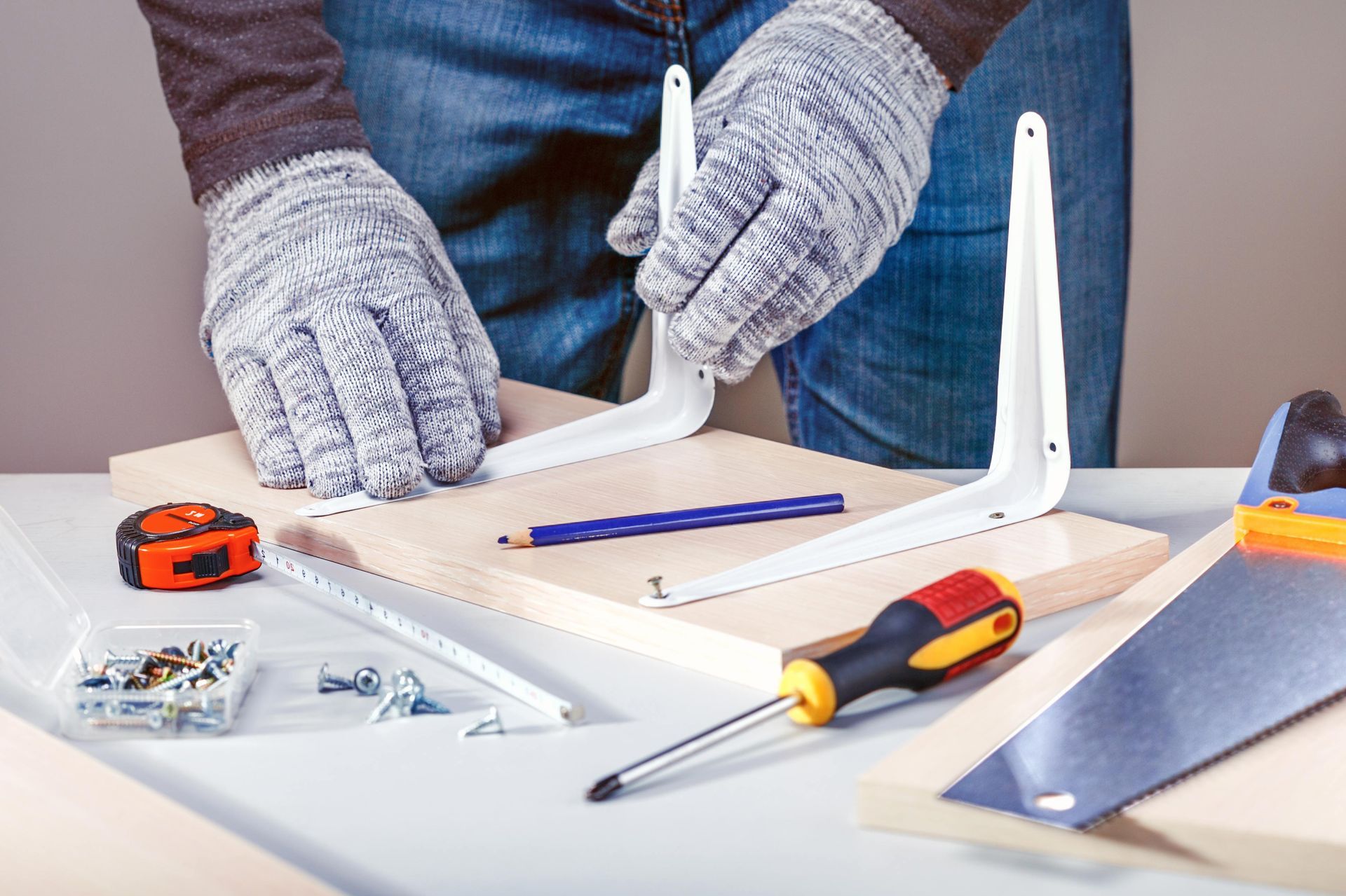 Gloved hands assembling a white furniture piece with screwdriver, screws, and tape measure on a table