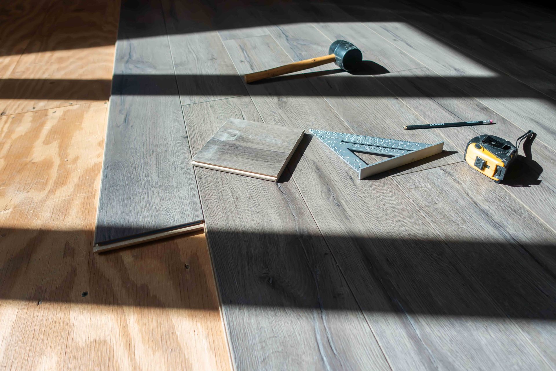 Gray floor tiles being installed on a sunlit floor, with tools and spacers nearby.