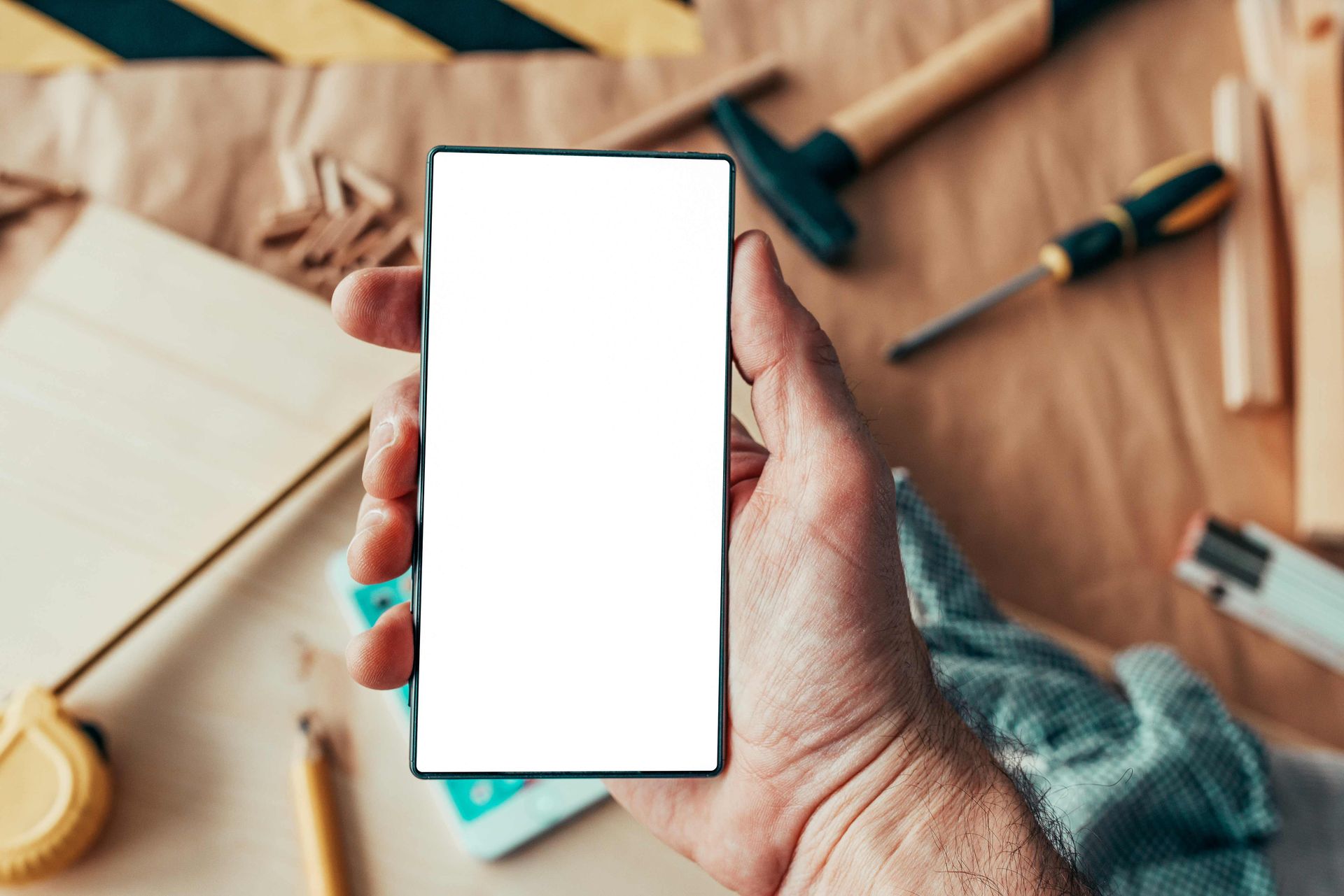 Hand holding a smartphone with a blank white screen over a desk with notebook and stationery