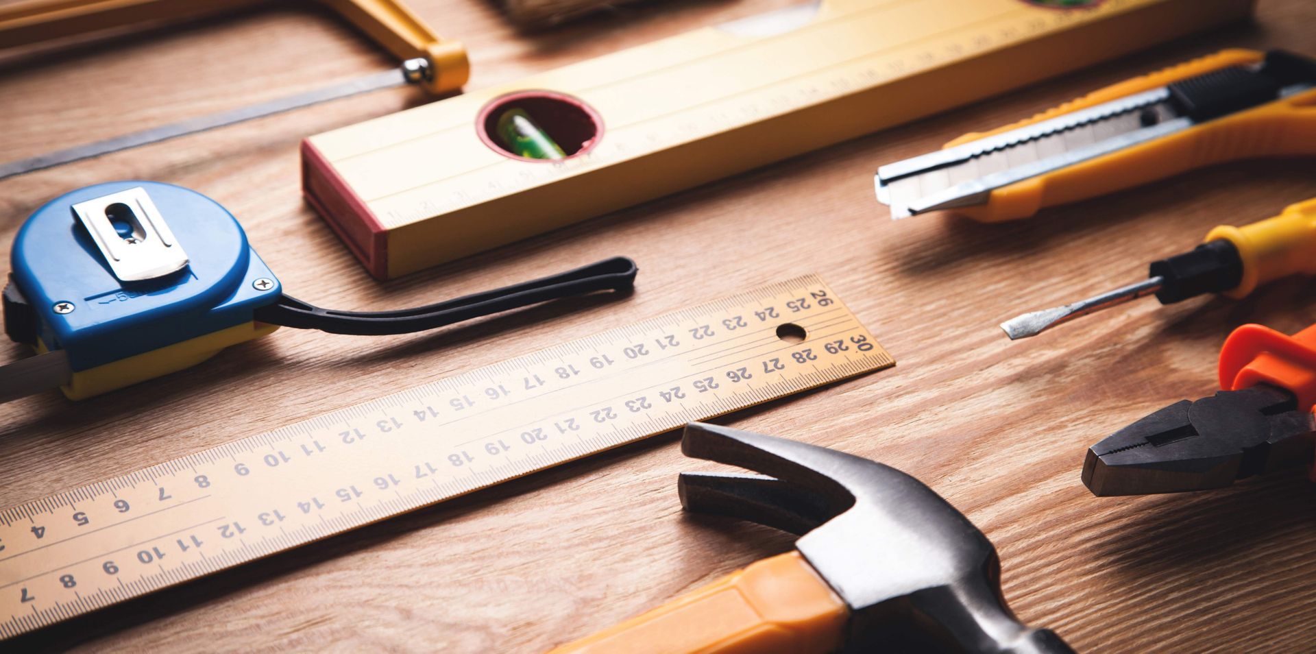 Assorted carpentry tools on a wooden workbench, including tape measures, ruler, saw, pencil, and hammer.