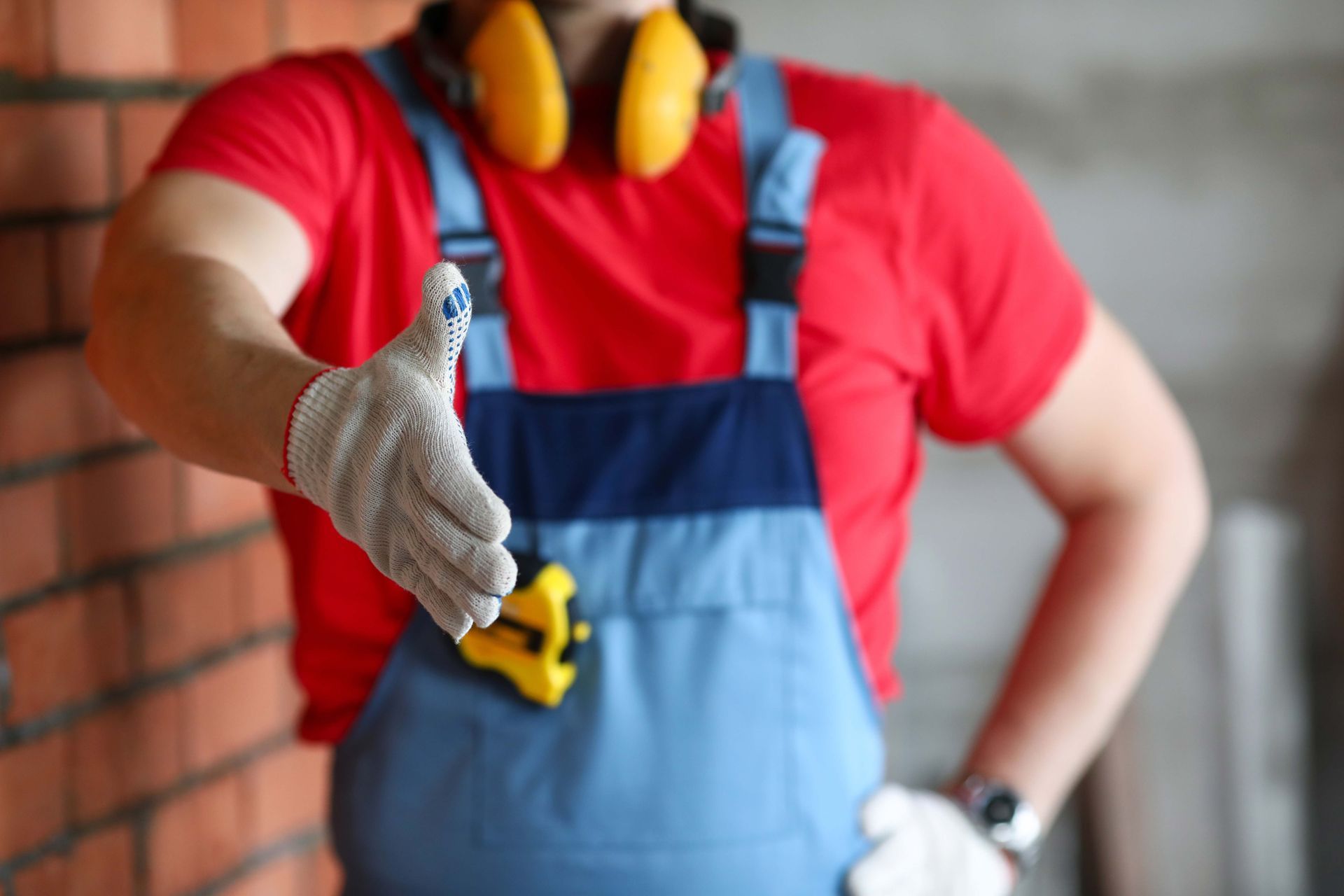 Construction worker in red shirt and blue overalls holding a yellow tape measure, giving a thumbs-up