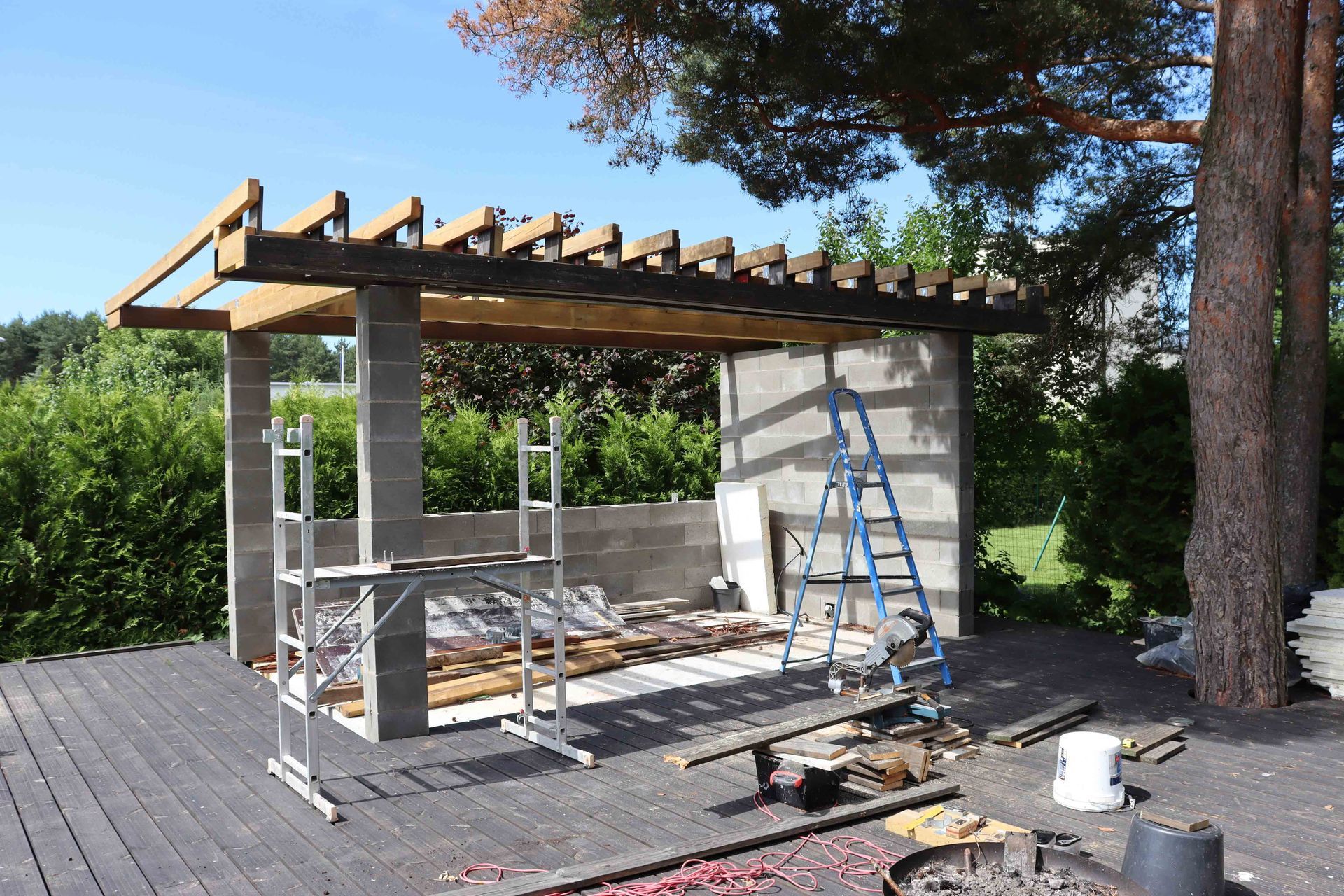 Partially built wooden pergola beside a concrete wall, with ladders and tools on a sunny deck