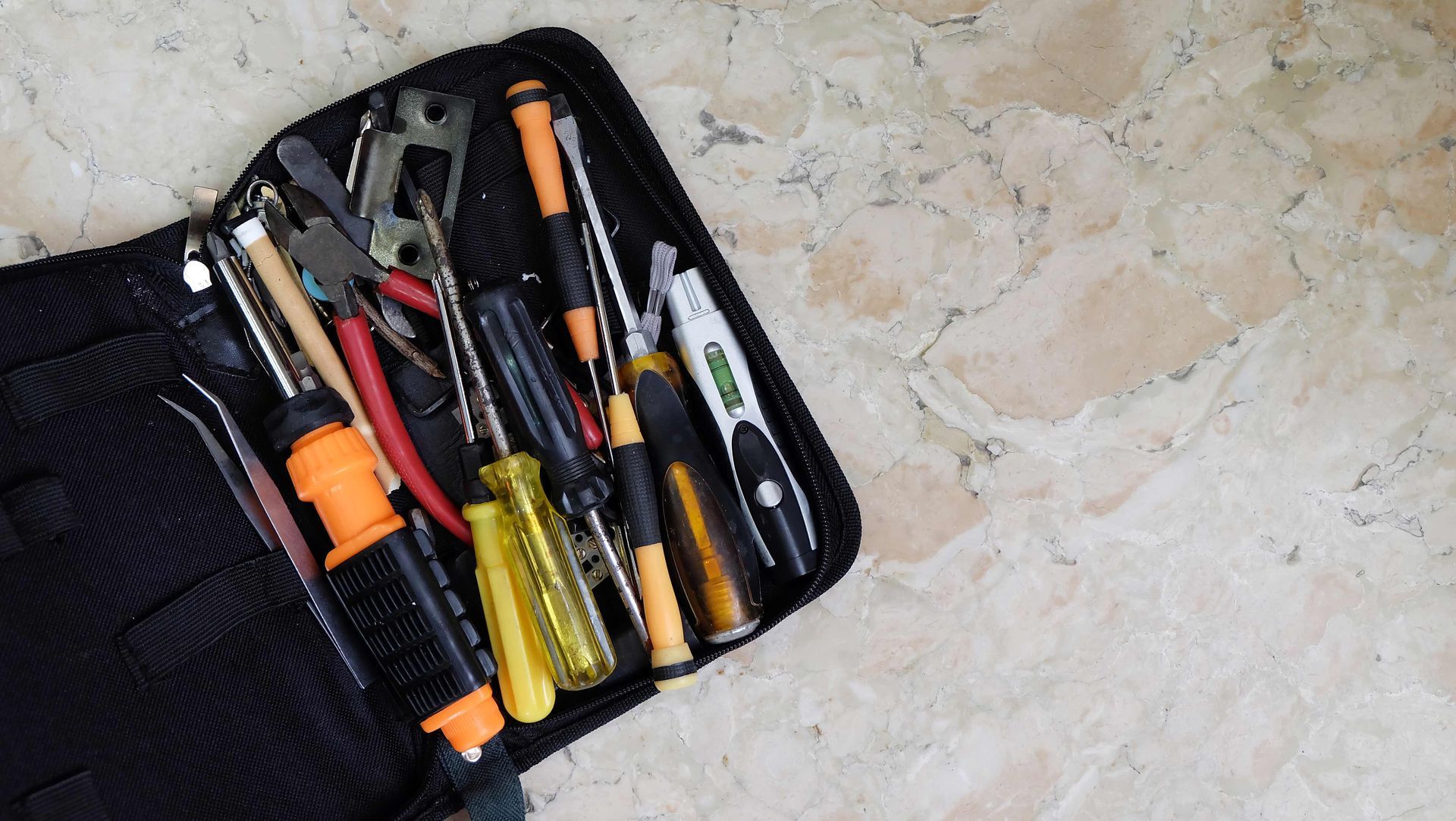 Open tool pouch with screwdrivers, pliers, and other hand tools on a light stone surface