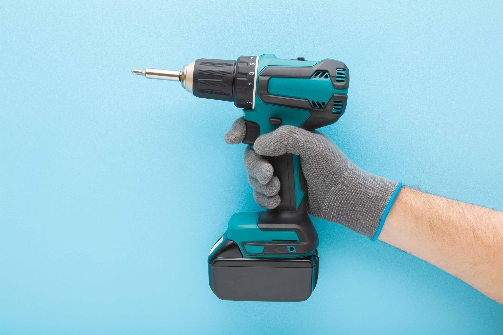 Gloved hand holding a teal cordless drill against a blue background