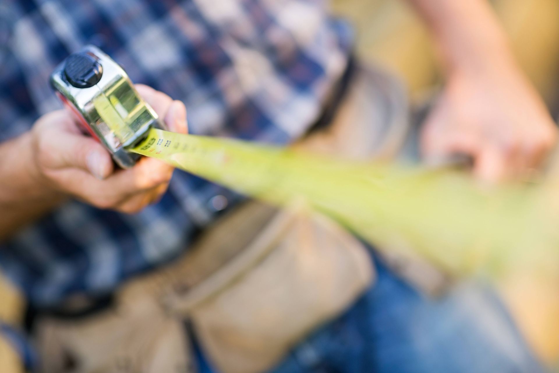 Measuring tape extending from a pocket tape measure held over a person’s jeans and plaid shirt