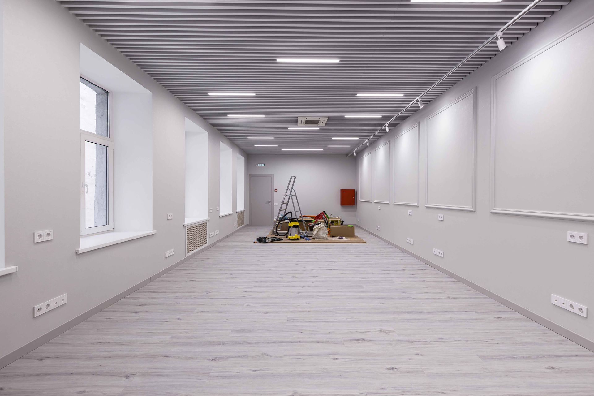 Empty white room with a gray floor, ceiling lights, and construction materials stacked at the far end.