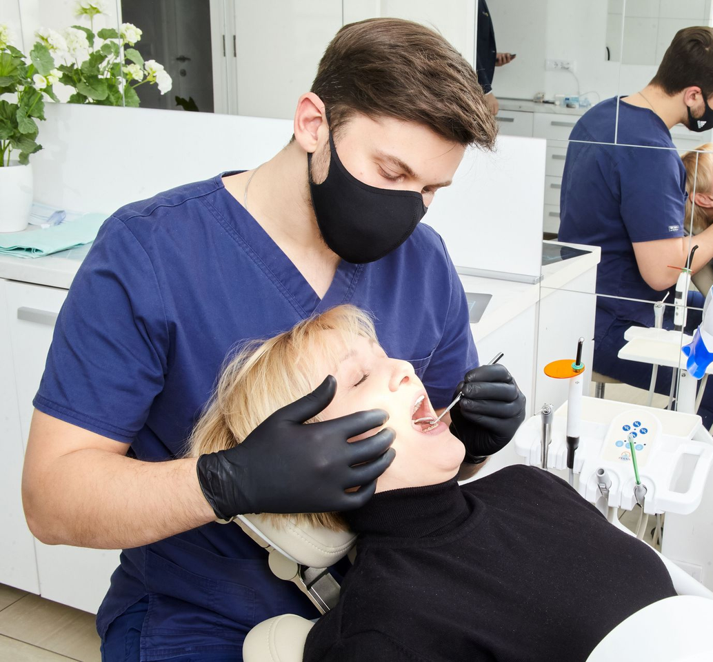 Dentist in blue scrubs and black mask examining a patient in a dental chair at a clinic