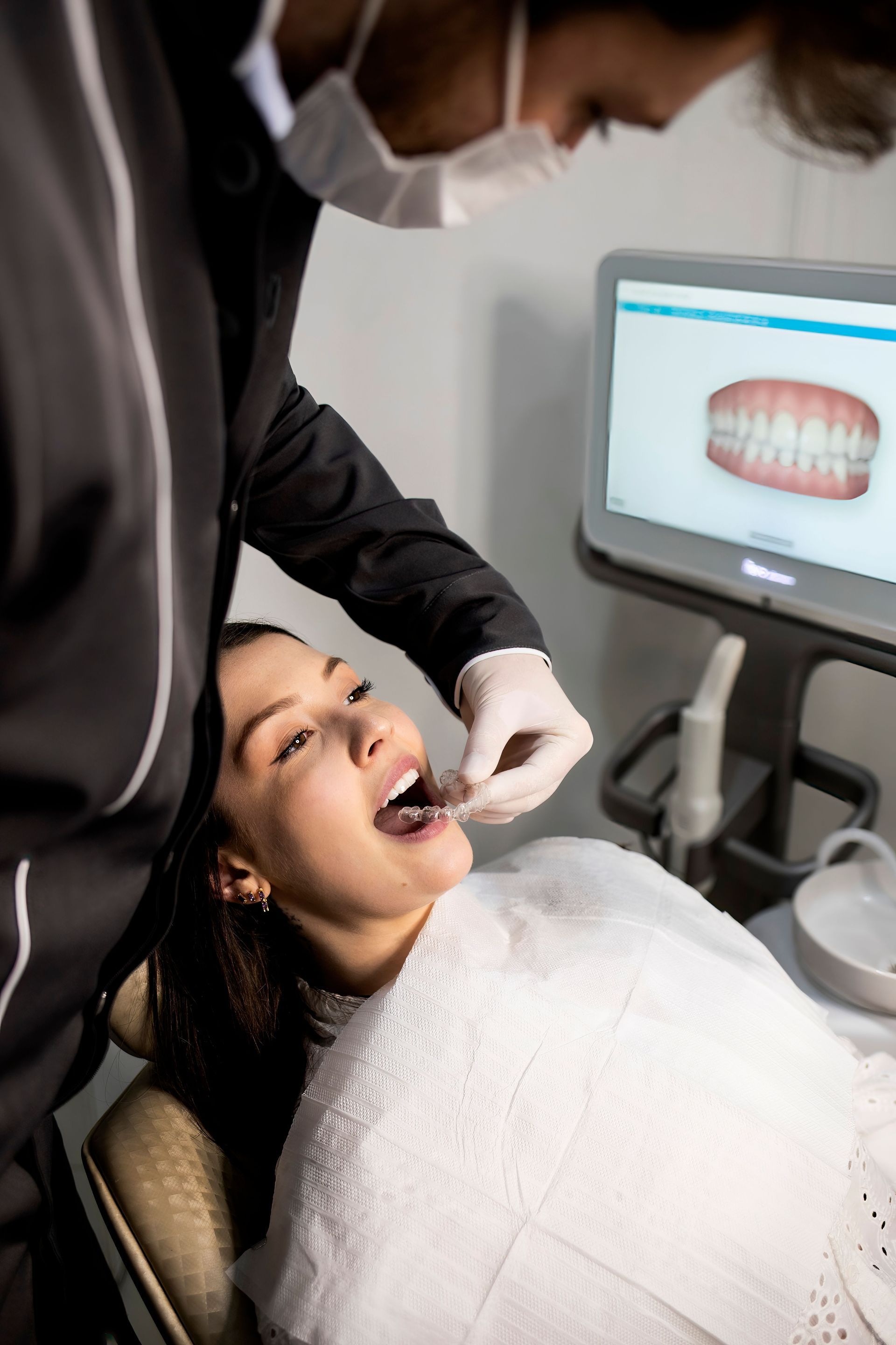 Dentist examining a patient’s mouth beside a monitor showing dental X-rays