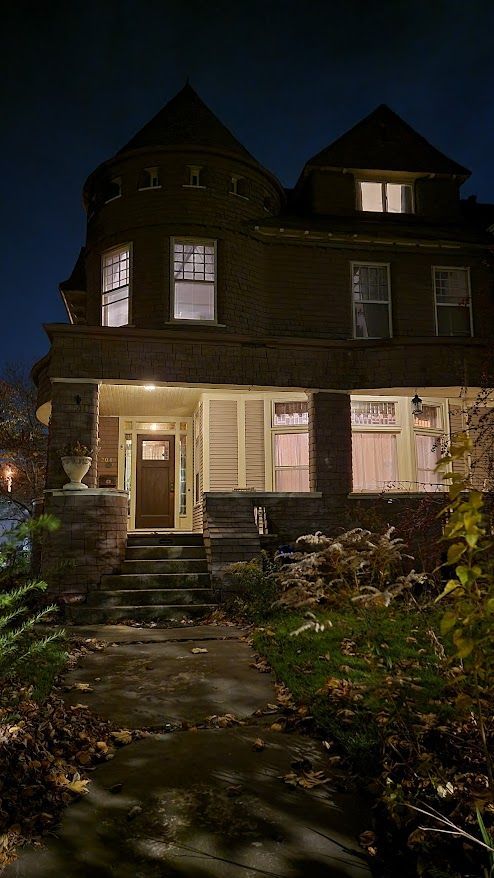A three-story house at night with lights on, steps leading to a porch, and a turret.