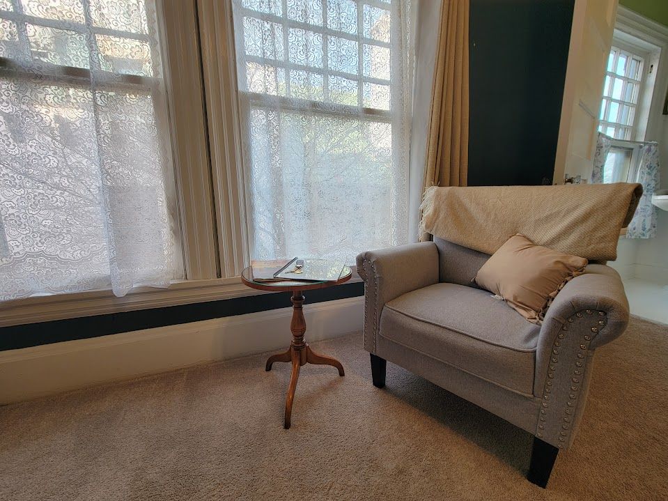 Cozy reading nook: gray armchair, small table, sheer curtains, window with sunlight.