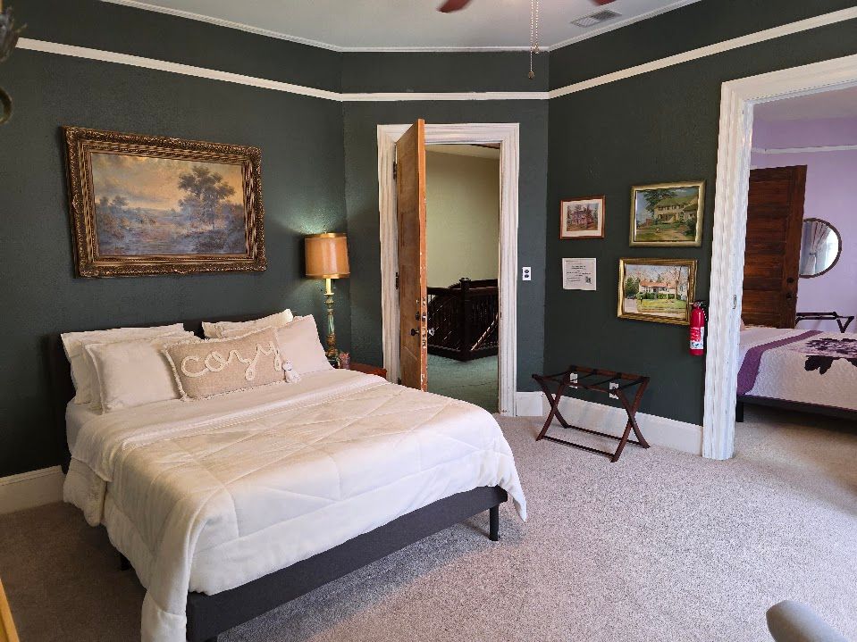 Bedroom with a bed, artwork, and dark green walls. Another room is visible through a doorway.