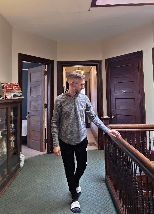 Man walking down a hallway, holding a railing. Gray and black clothing, interior, and wood doors.