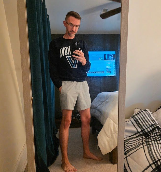 Man in glasses and Villanova sweatshirt taking mirror selfie in bedroom, wearing shorts.