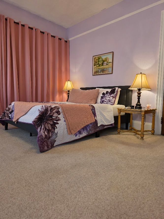 Bedroom with bed, nightstand, lamps, and pink curtains. Purple walls and carpet.