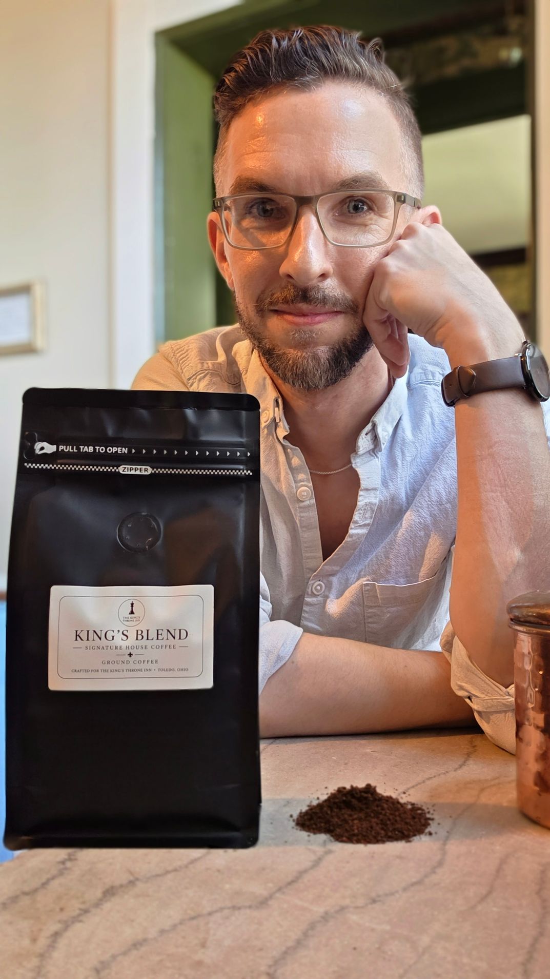 Man smiling behind a black coffee bag on a table, with coffee grounds and a mug nearby