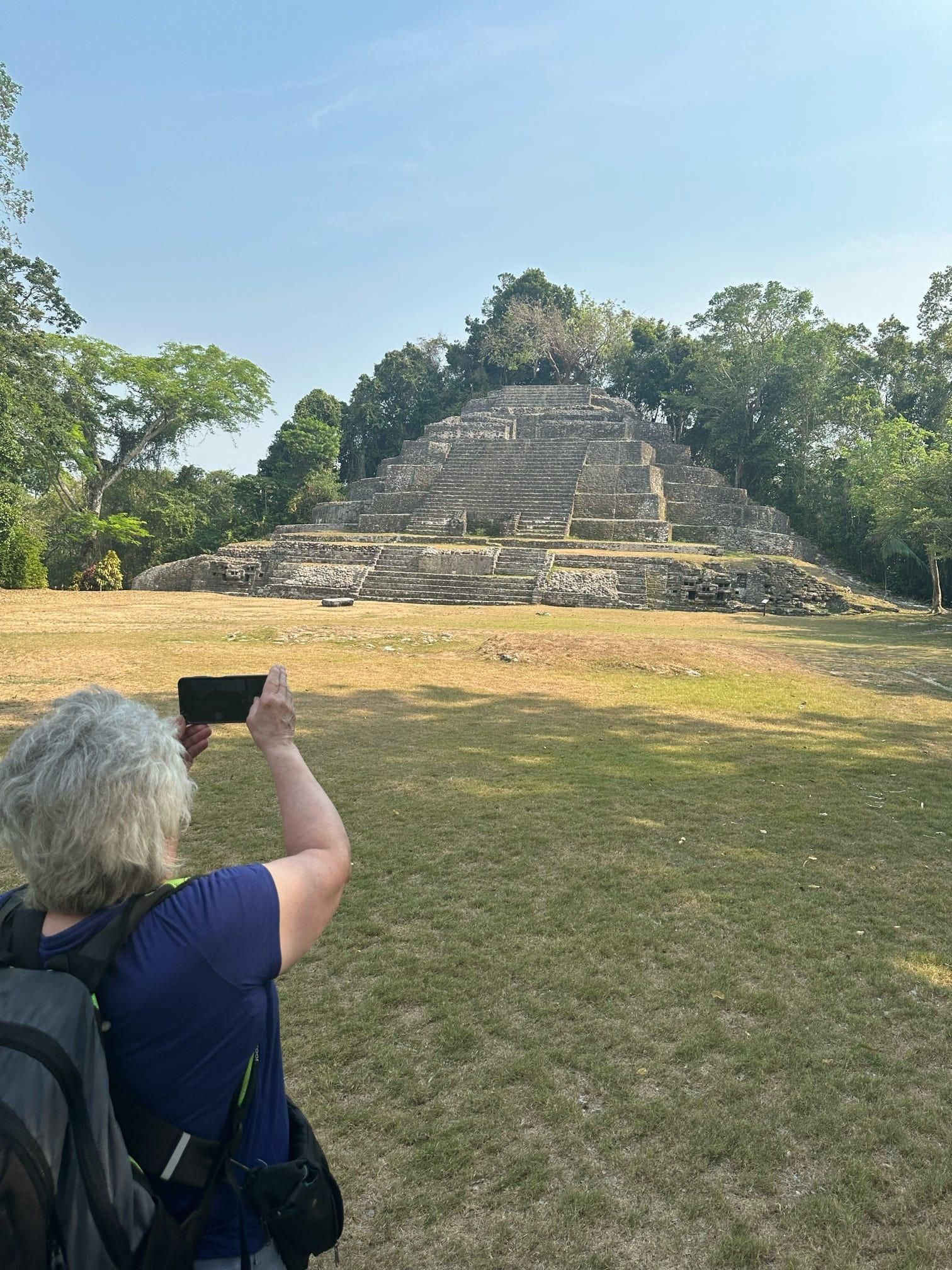 Person taking a photo of an ancient stone pyramid in a grassy, tree-lined archaeological site