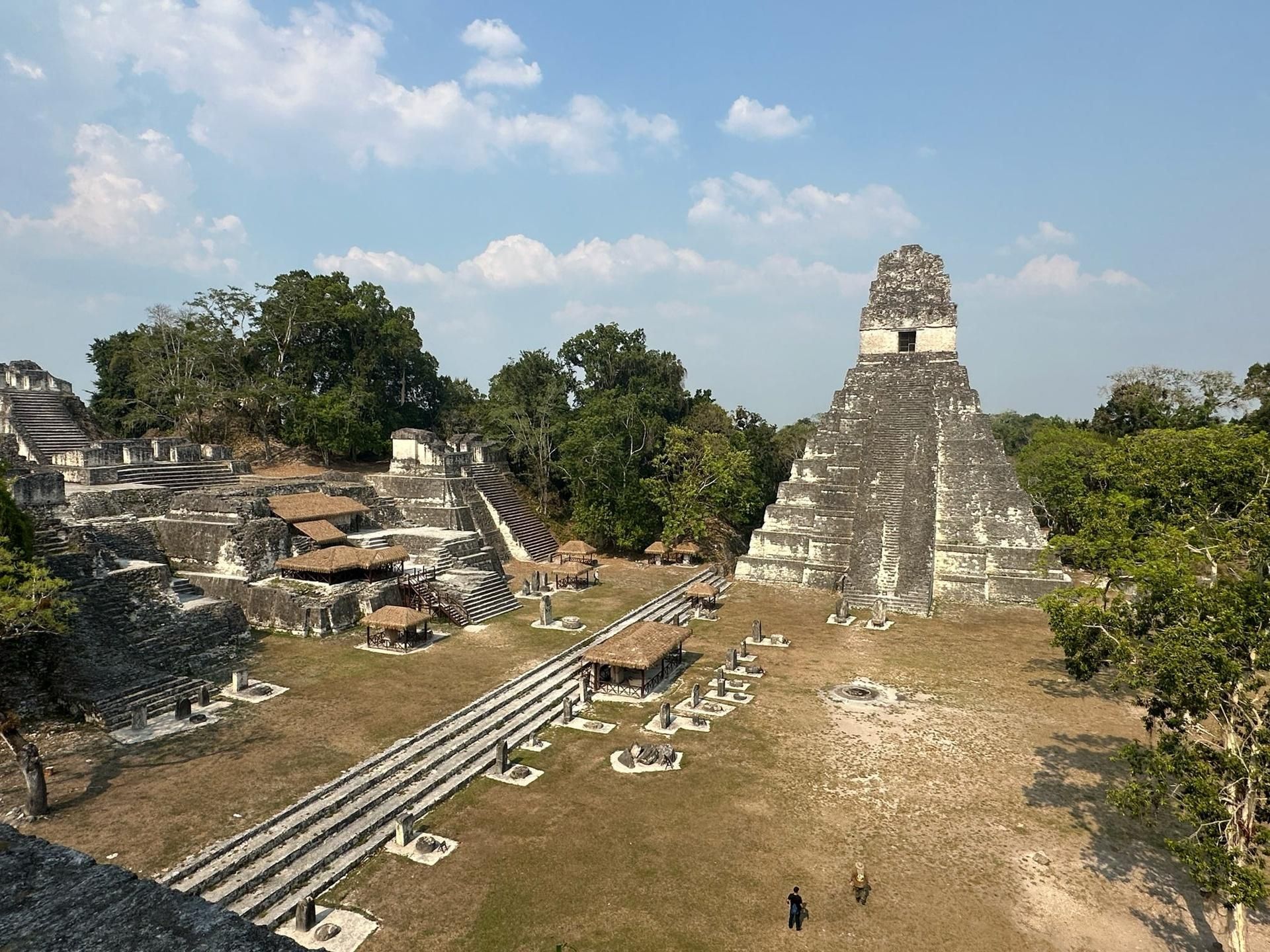 Ancient stone pyramid ruins in a grassy archaeological site under a blue sky, with trees and pathways nearby