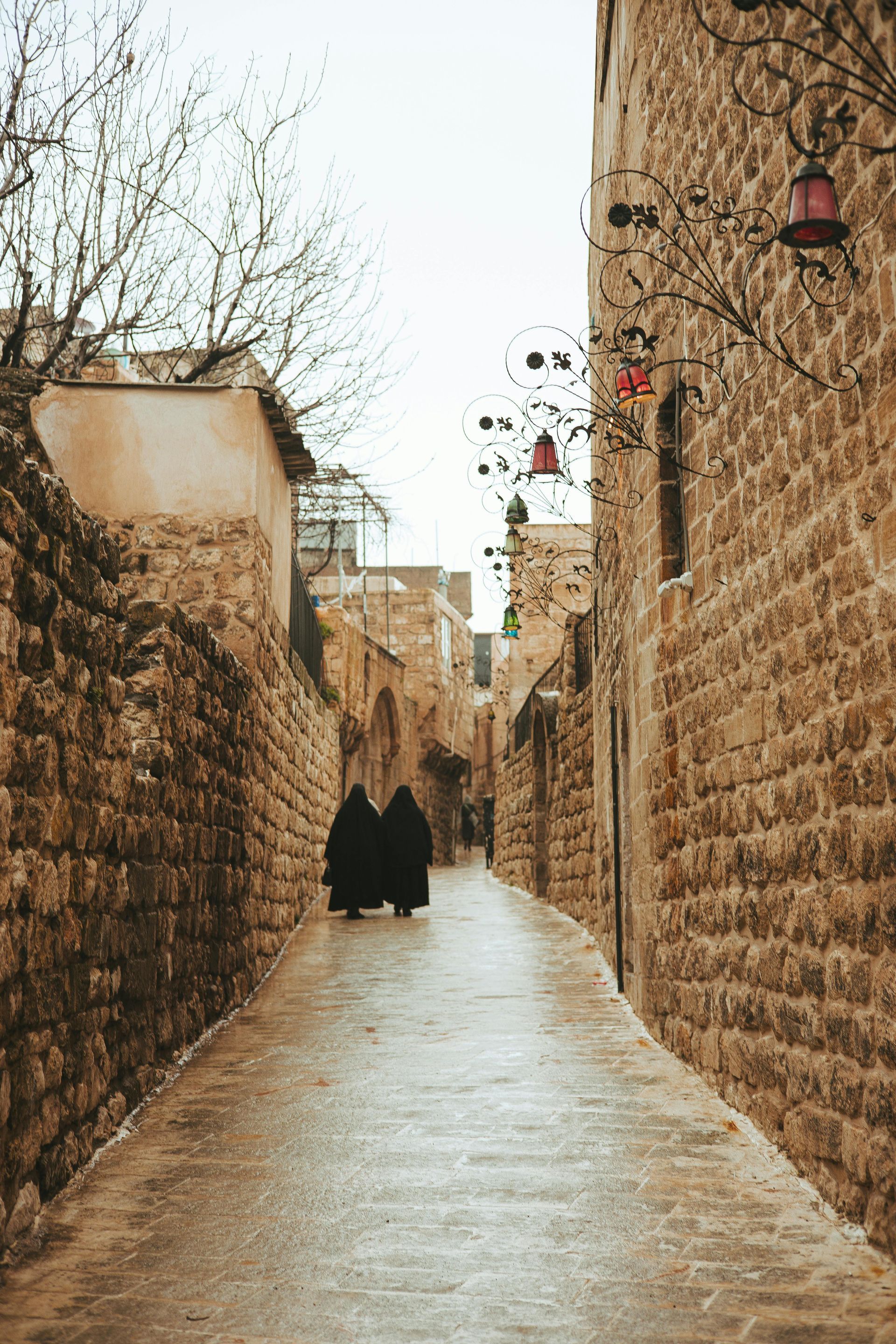 Narrow stone alley with wet pavement and two people walking away under bare trees and hanging lanterns