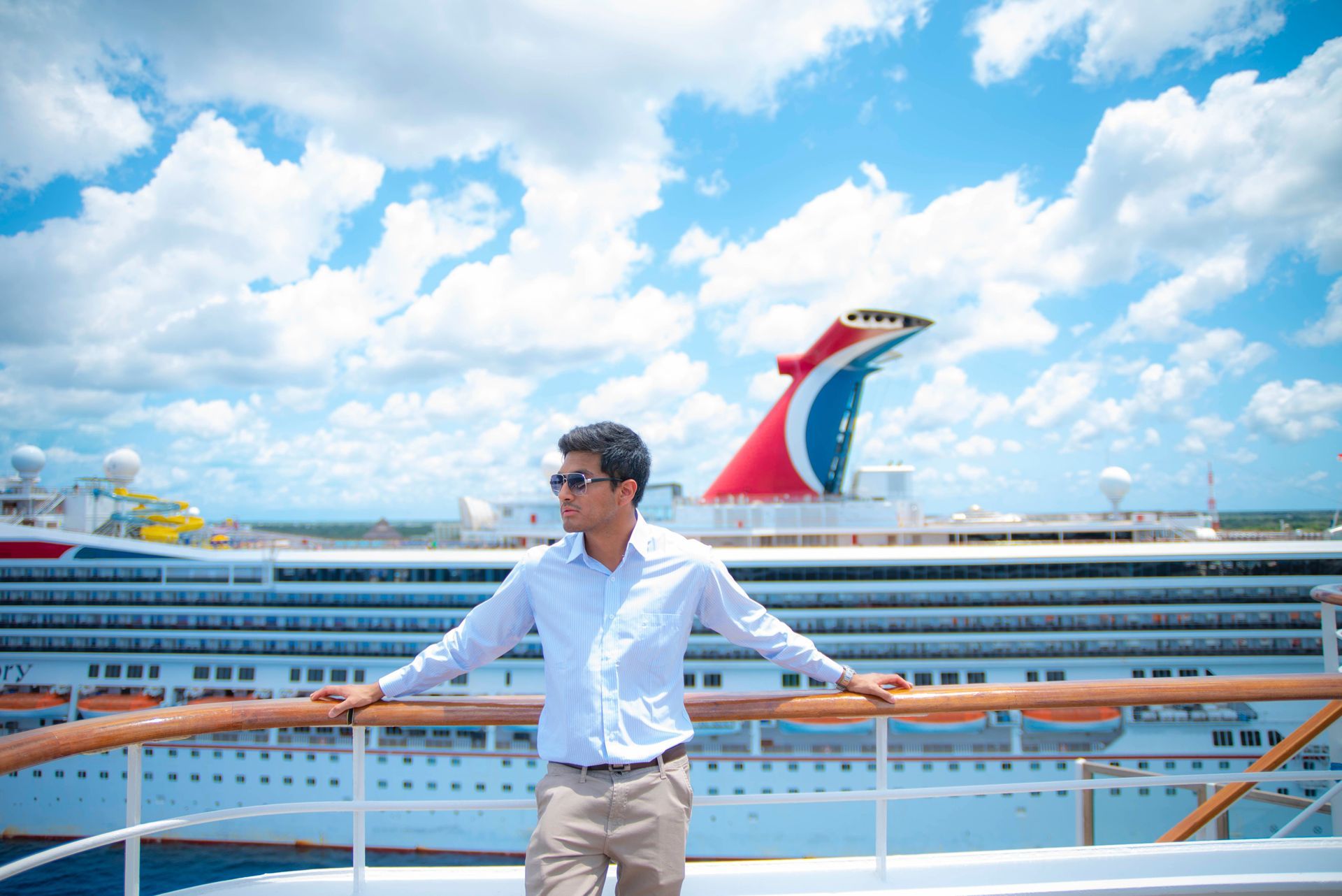 Man standing on a cruise ship deck under a bright blue sky with a red-and-blue funnel in the background