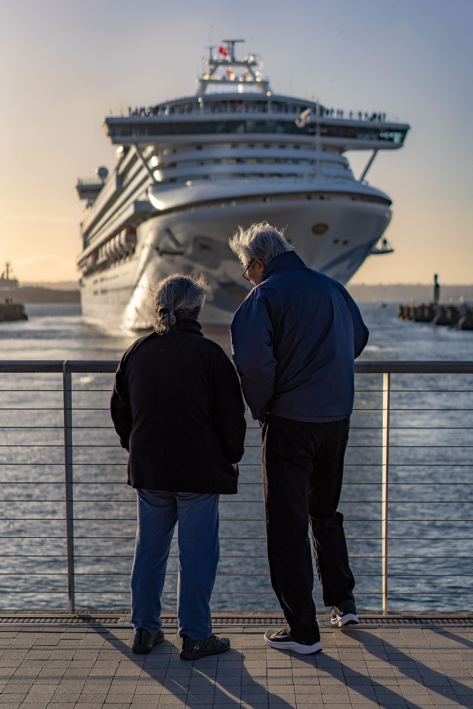 Two people stand on a waterfront railing watching a large cruise ship approach at sunset.