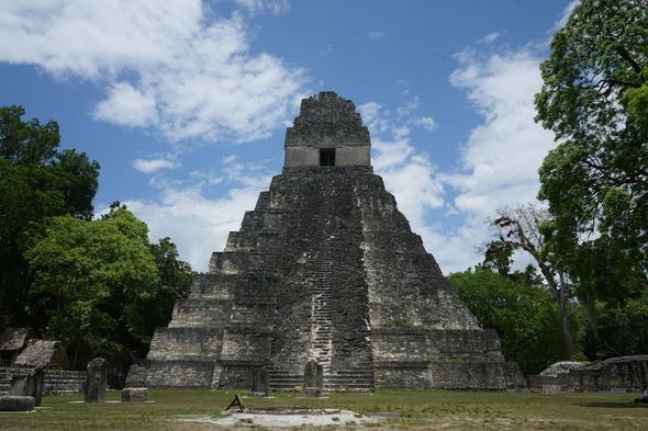 Ancient stone Mayan pyramid under a blue sky, surrounded by trees and grass