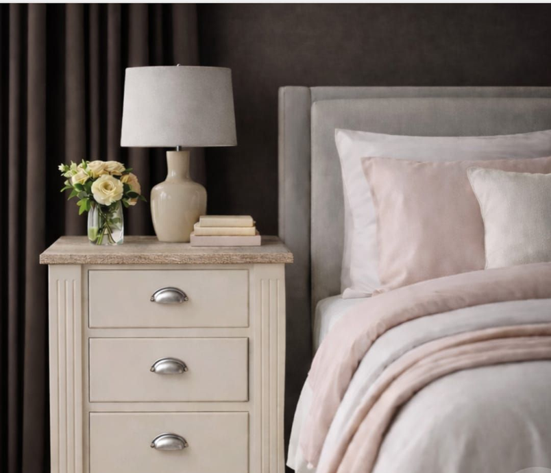 Mesita de noche con lámpara, flores y libros junto a una cama con sábanas de color rosa suave.