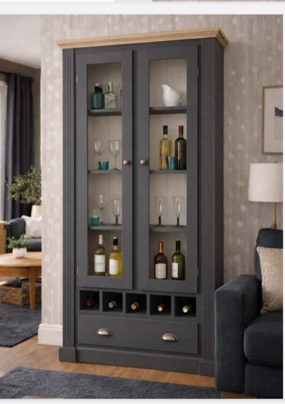 Vitrina gris con puertas de cristal y estantes en una sala de estar, que contiene botellas y elementos decorativos.