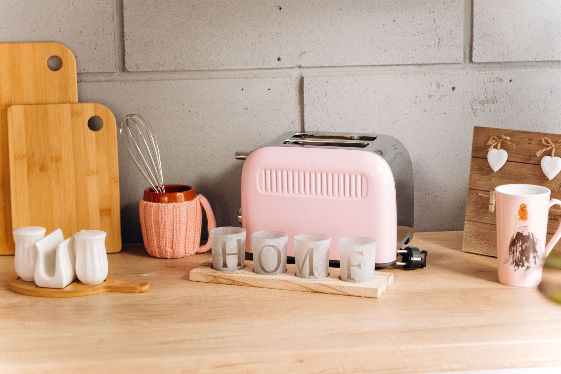 Tostadora rosa y tazas sobre una encimera de madera en la cocina, junto a una pared de ladrillo claro.