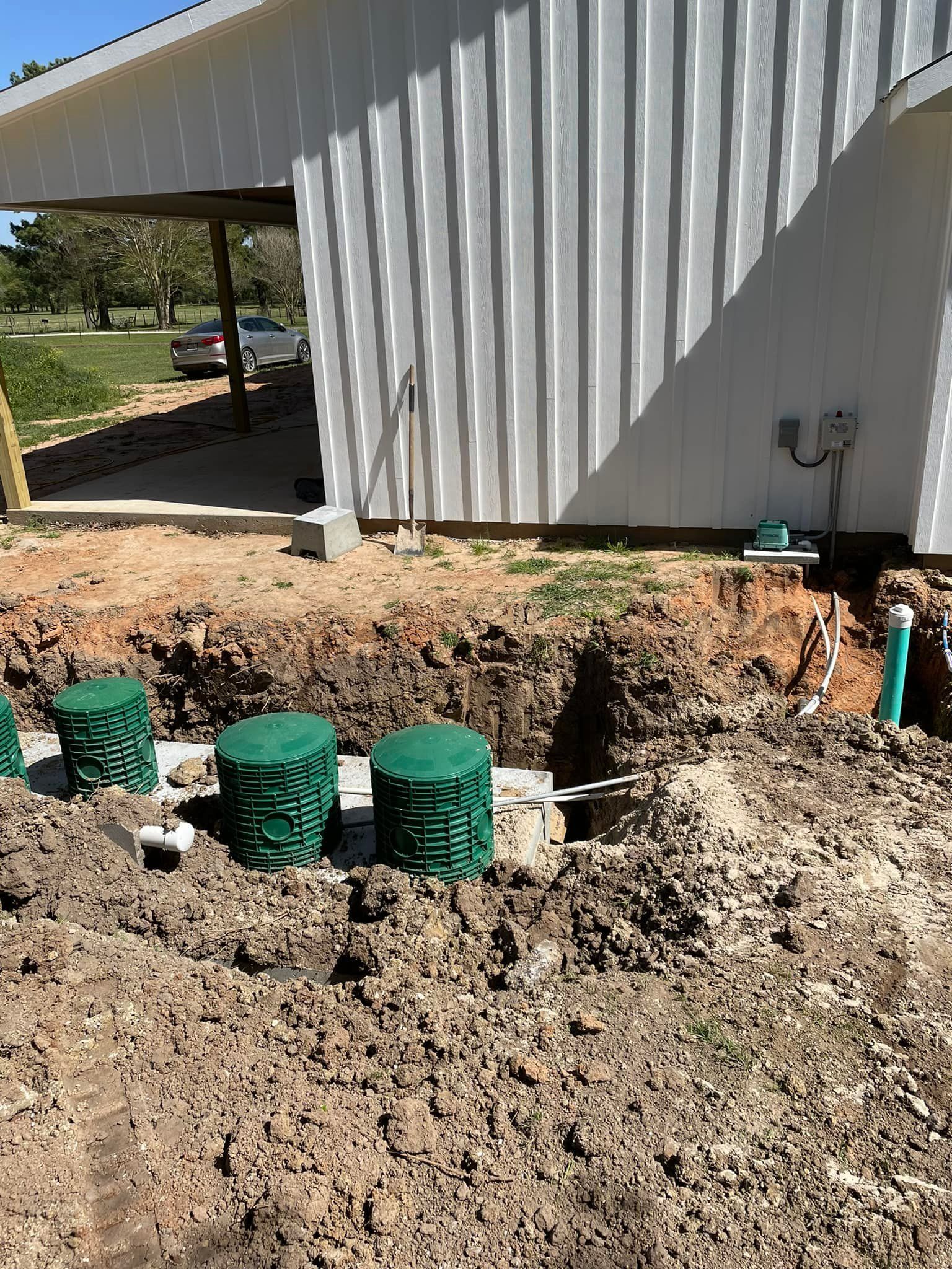 Trenches around a house foundation with green utility pipes and exposed dirt in front of a metal-sided building