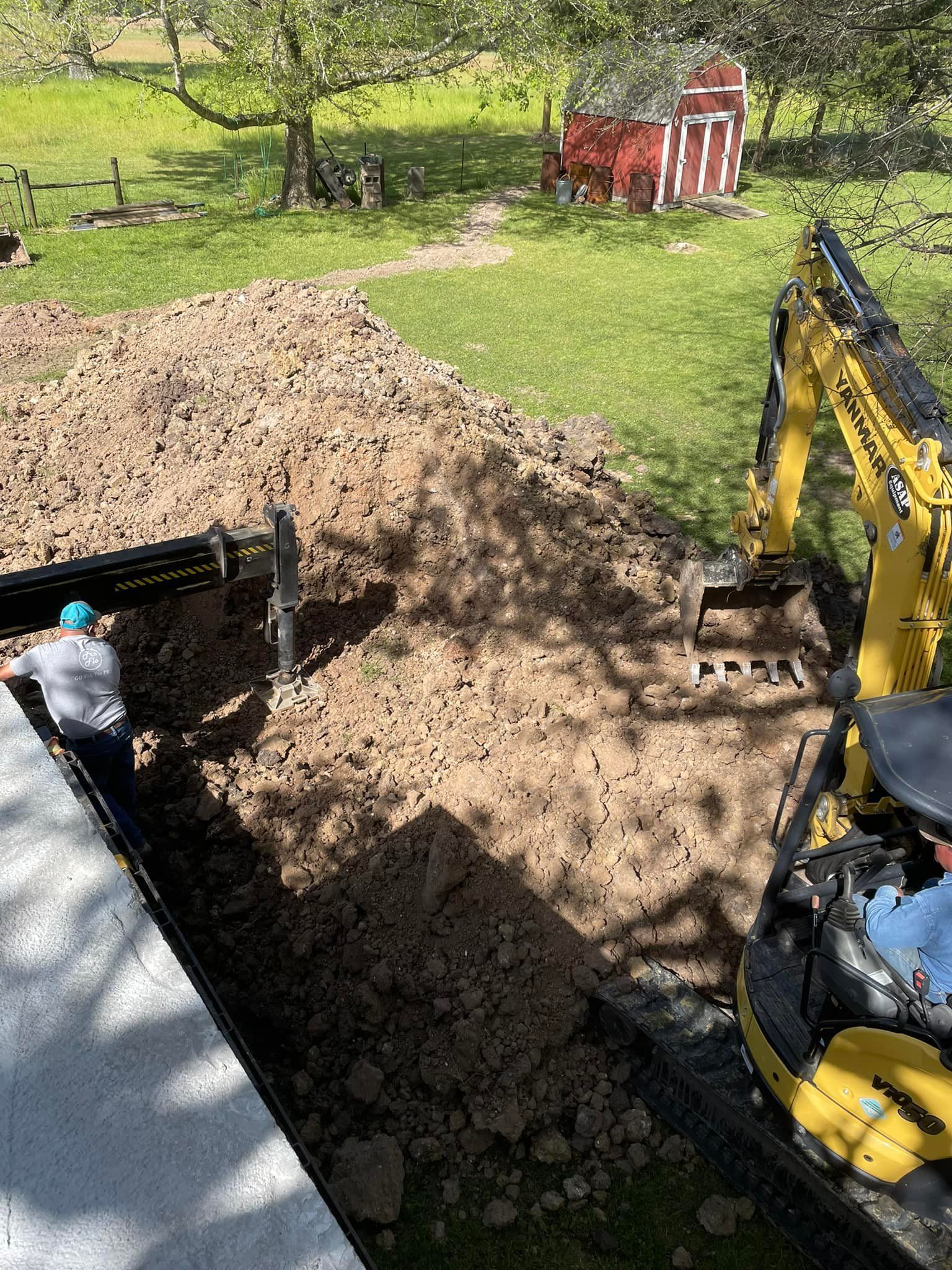 Yellow excavator beside a large dirt pile in a backyard, with a worker near the house and a red shed in the background
