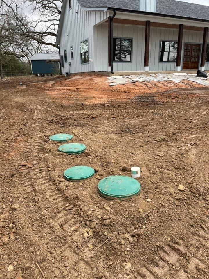 Four green utility covers in a dirt yard beside a gray house with porch columns.