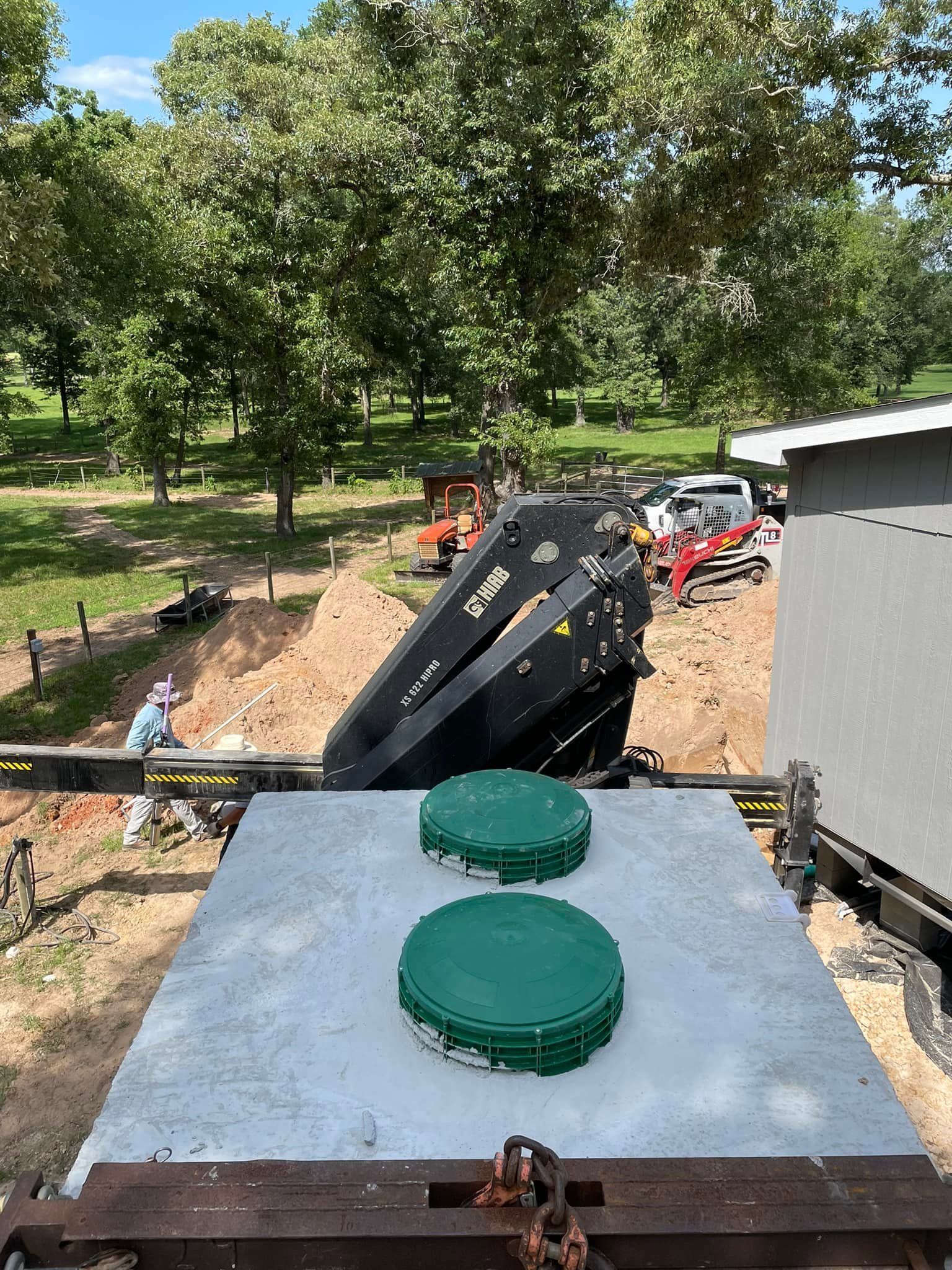 Two green tank lids on a metal platform overlooking a backfilled trench with an excavator in a wooded yard