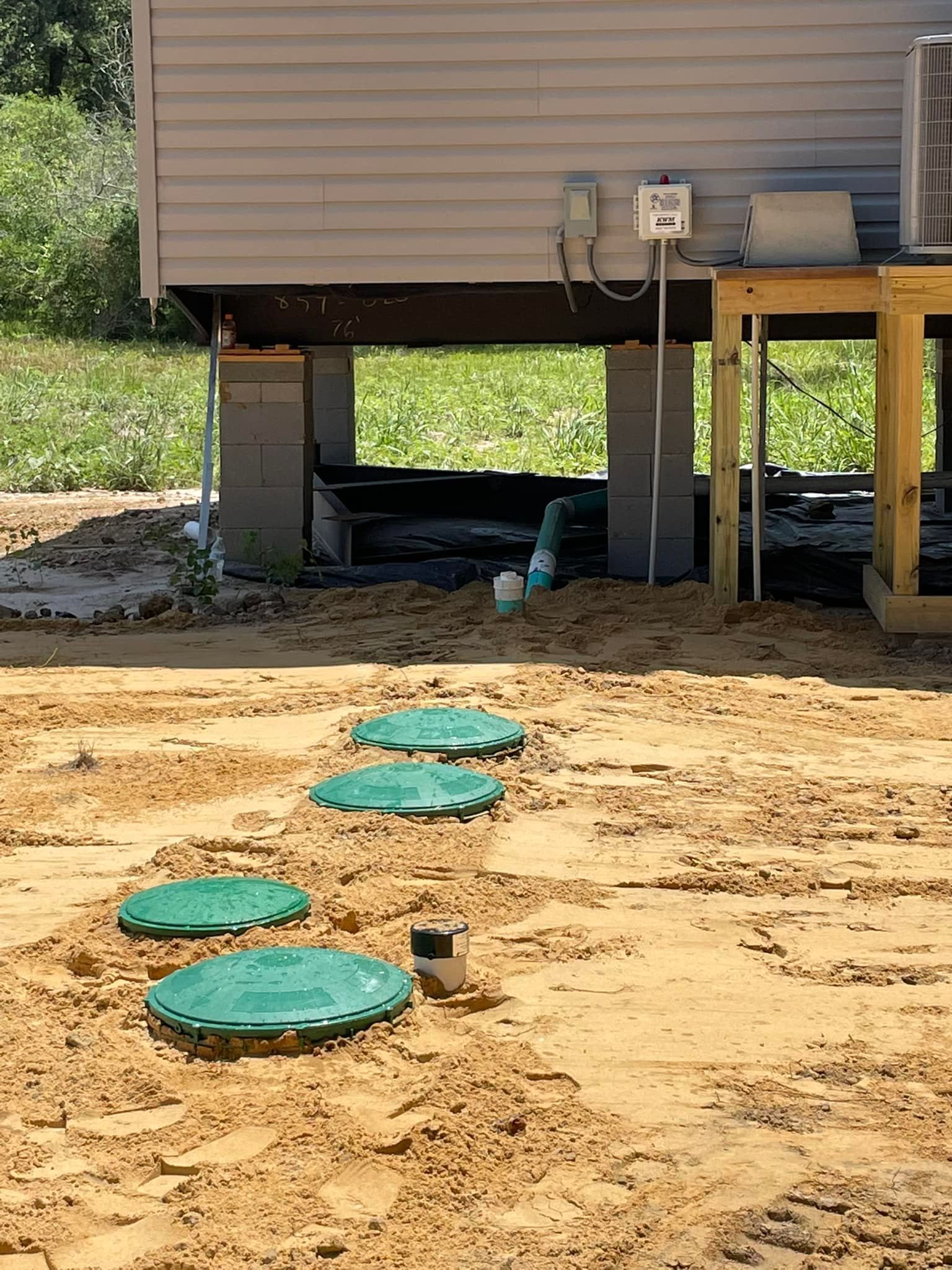 Crawlspace under a house with utility pipes and green septic lids in sandy ground.