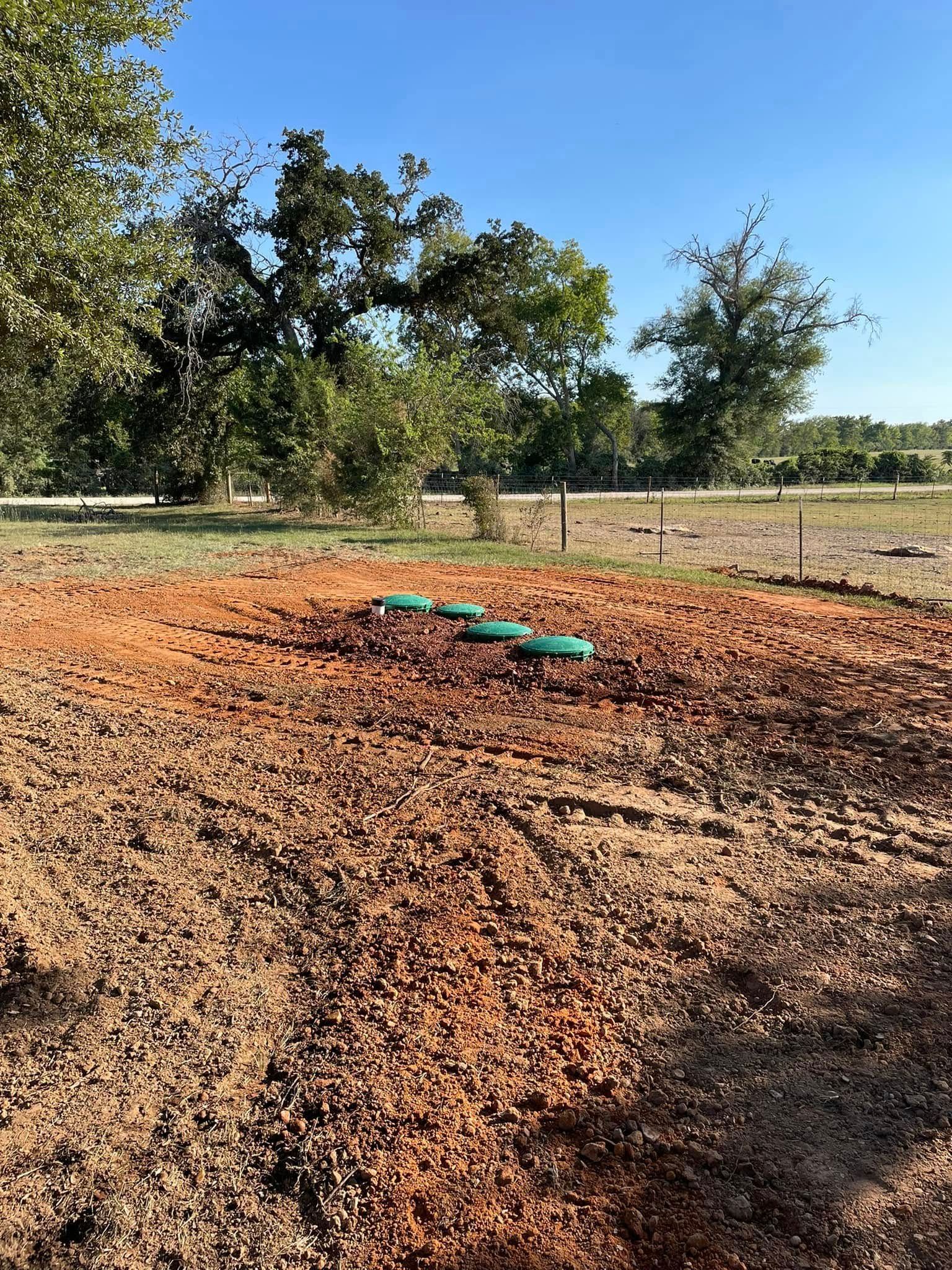 Rural dirt lot with a green tarp-covered pile, trees, and a fenced field under a clear blue sky.