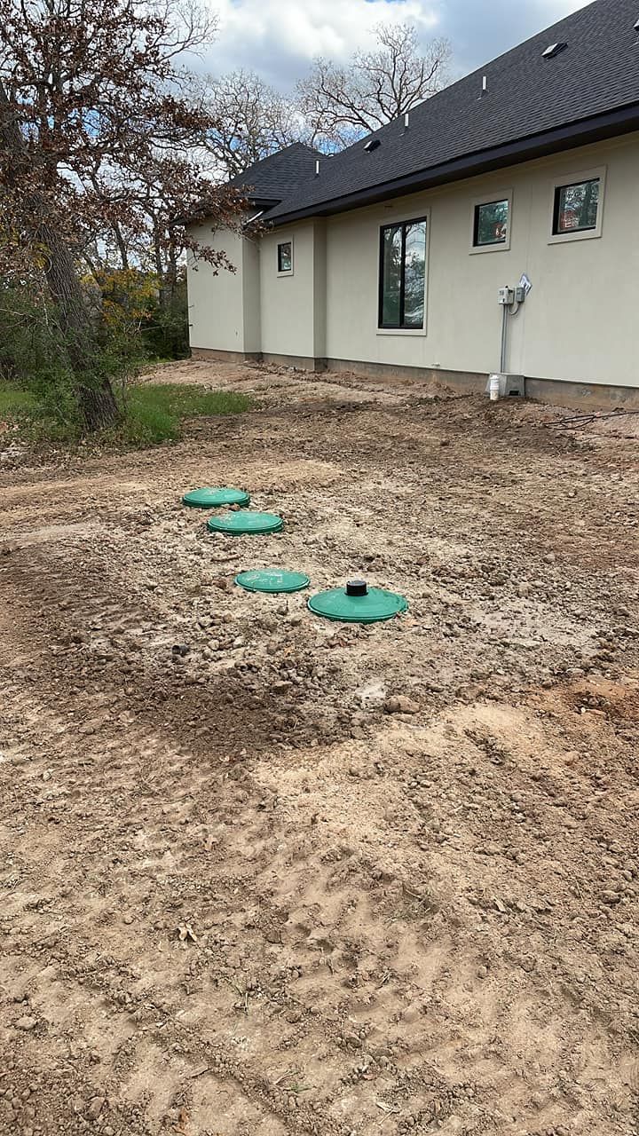 Three green utility covers in a muddy yard beside a house with a beige exterior and dark roof.
