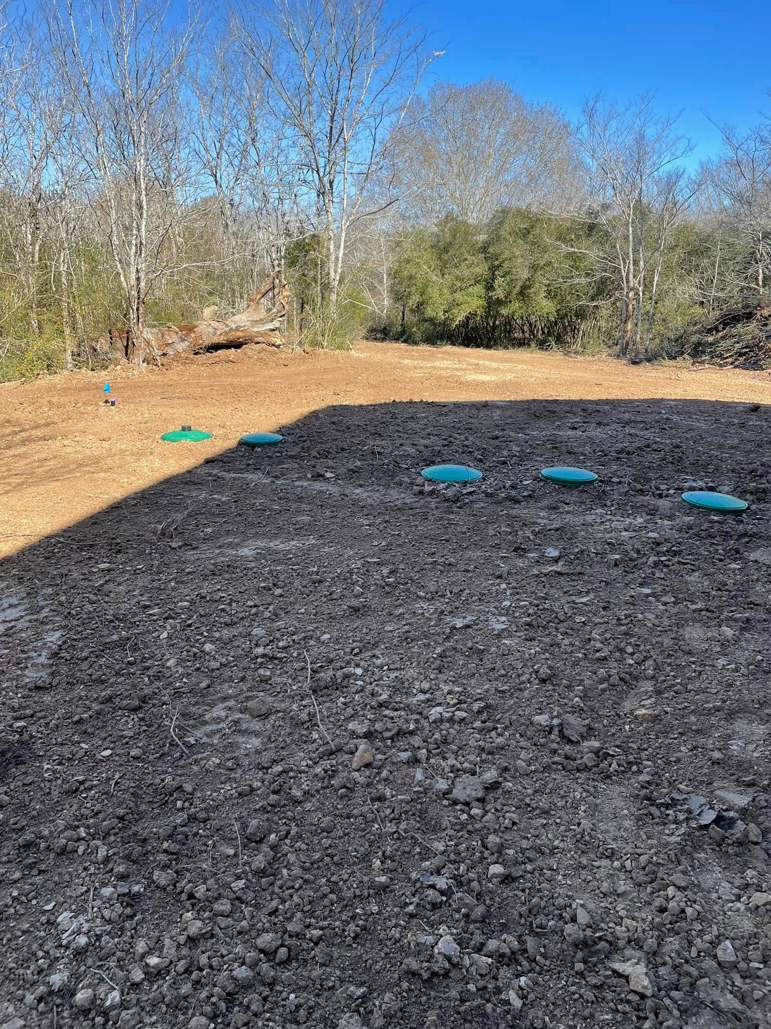 Gravel driveway beside a mulched yard with scattered blue markers and leafless trees in the background