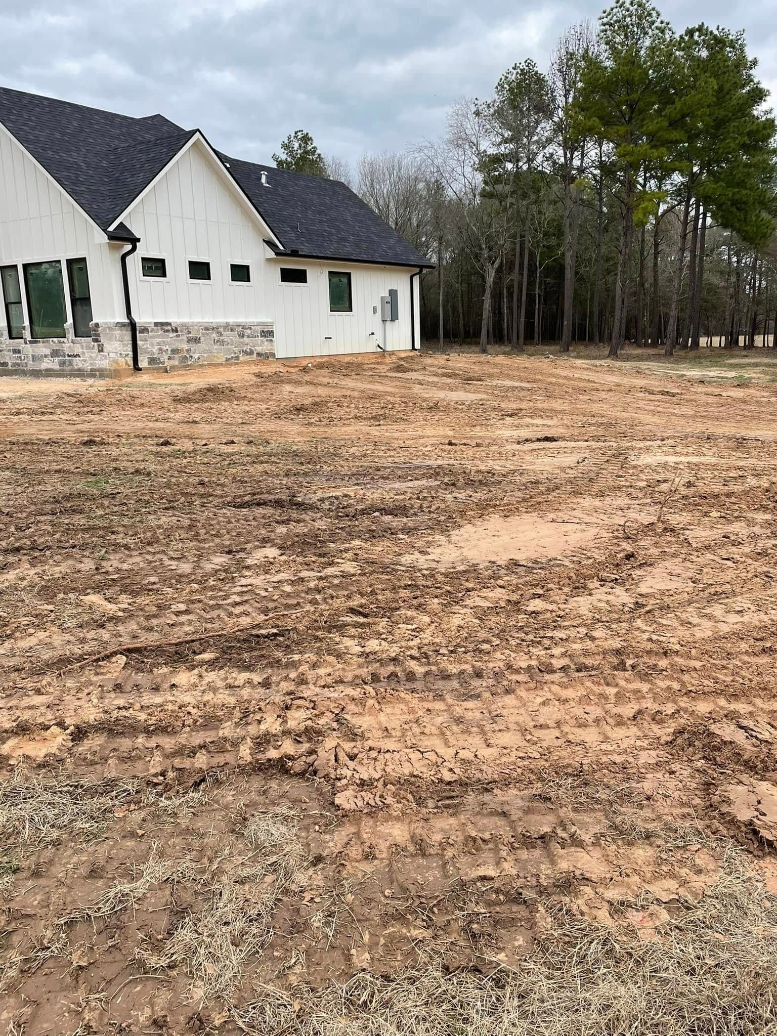 White house with dark roof beside a large muddy cleared lot and trees under an overcast sky