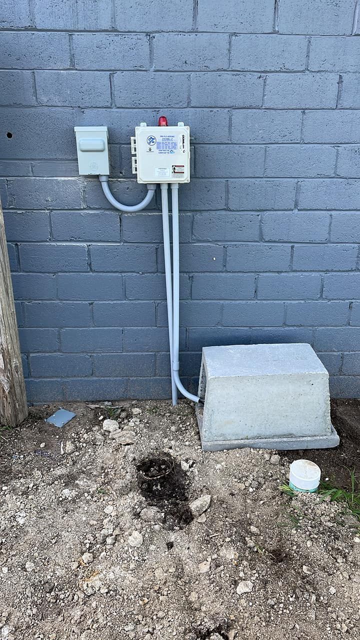 Electrical meter and conduit mounted on a gray cinderblock wall beside a concrete base on bare ground.