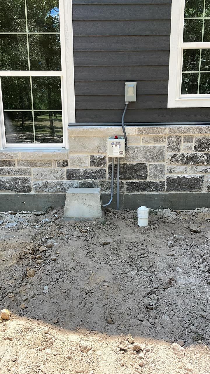 Exterior house wall with windows, electrical outlet, and utility boxes above a gravel ground area