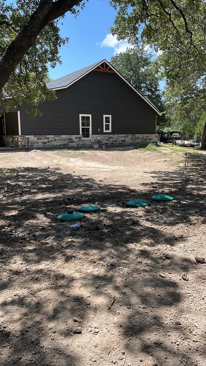 Dark building with stone base in a shaded dirt yard, with trees framing the scene and two teal circles on the ground