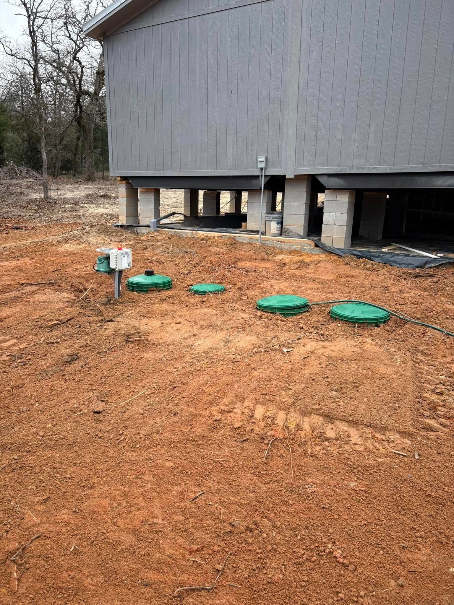Gray cylindrical building on stilts above a dirt yard with green utility covers and a small sign.