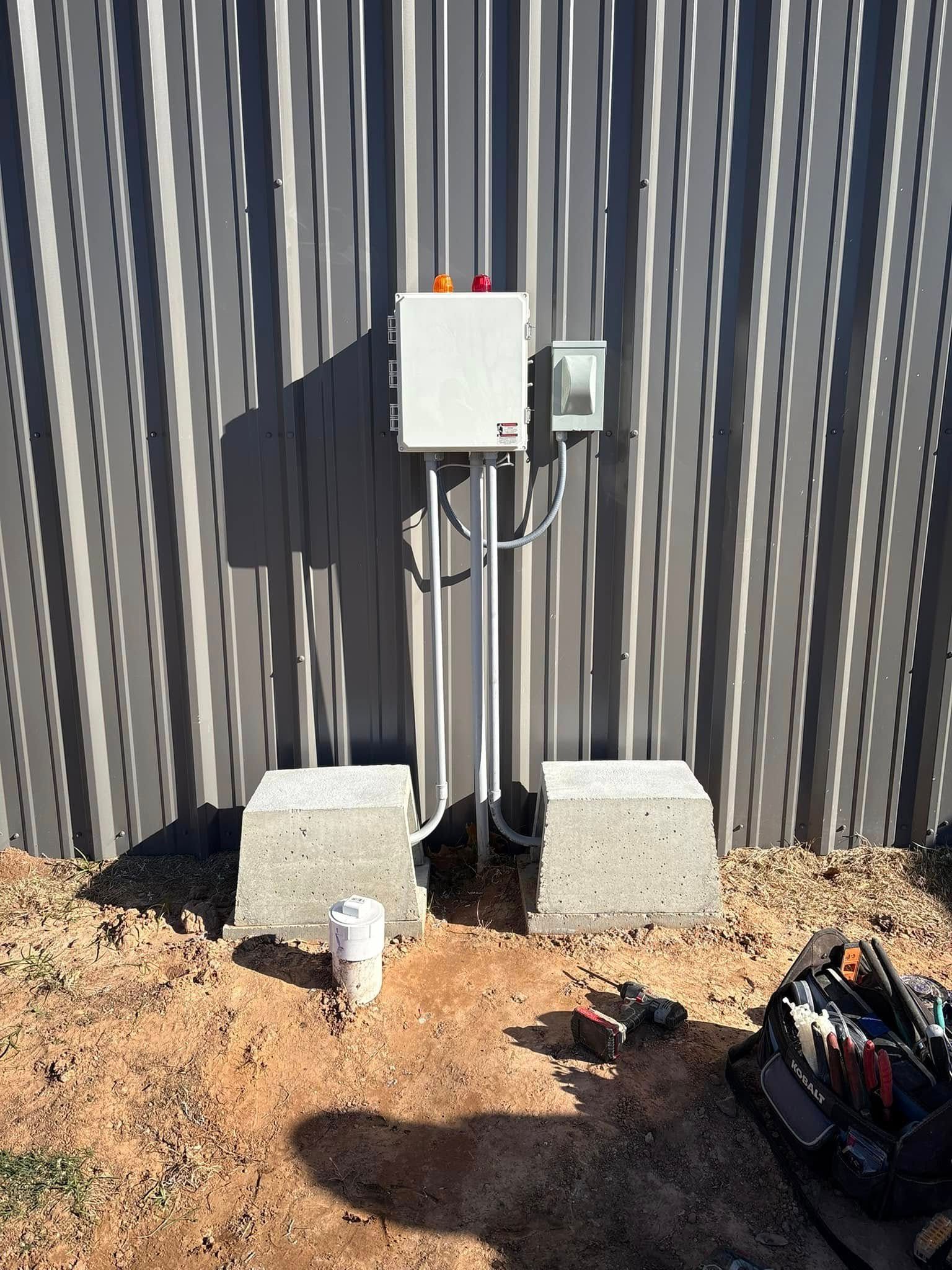Electrical control box on a striped wall, with concrete blocks and cables on a dirt ground.